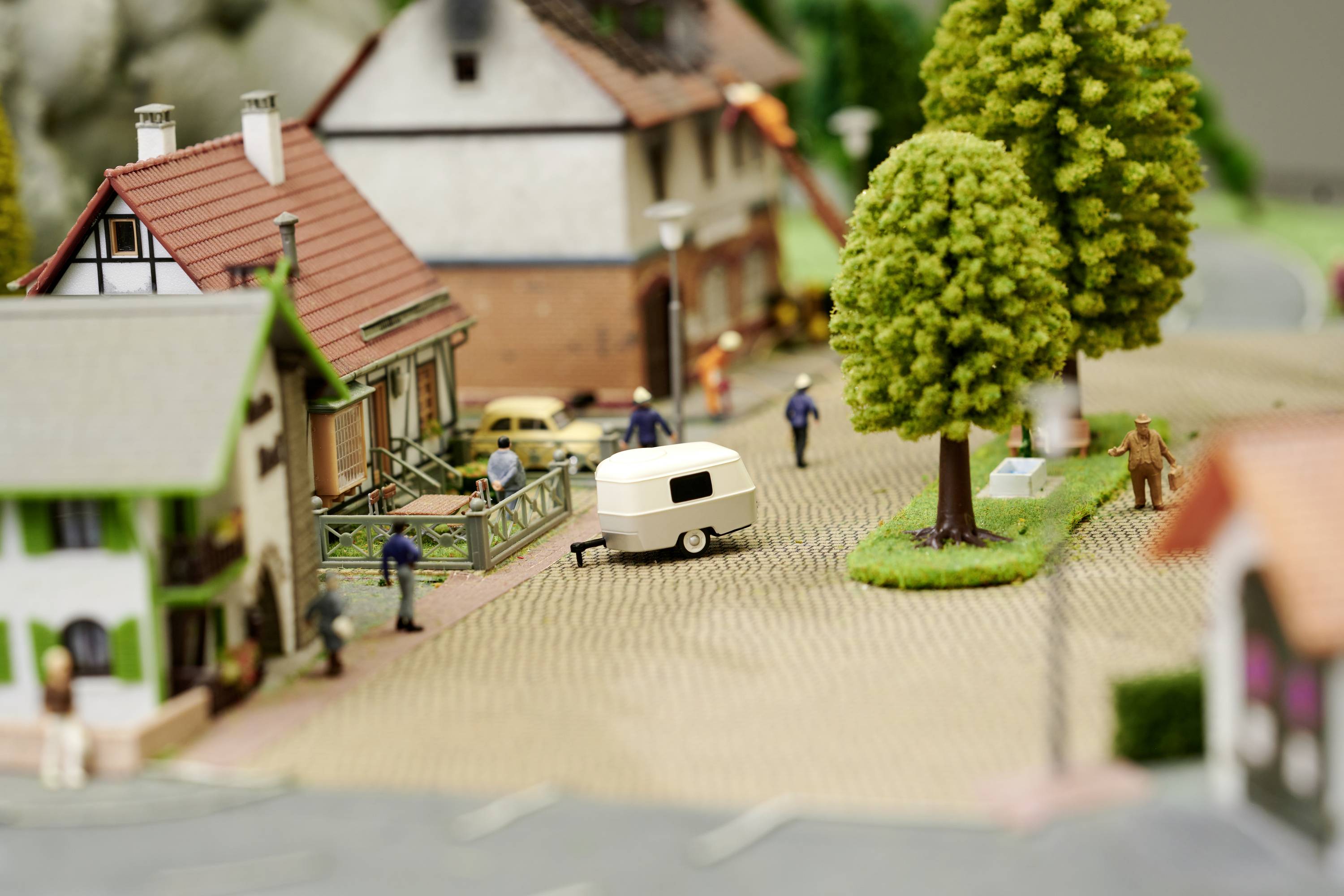 Miniature village with houses, trees, and figures. People standing close together, a caravan nearby. Scenic village street.