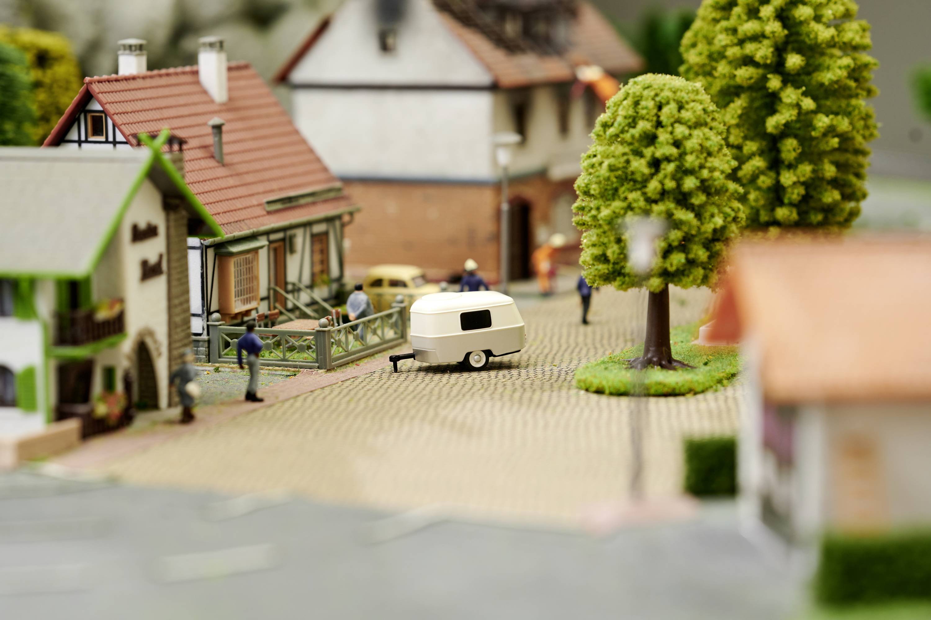 Miniature village scene with houses, figurines and trees. A caravan is parked on the street. Vivid details convey rural life.