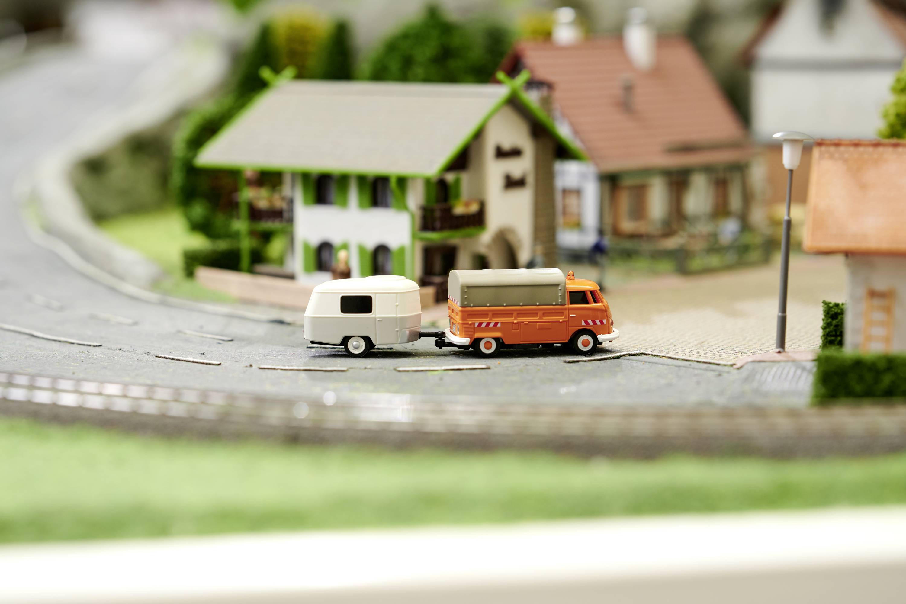 A miniature model depicts an orange van with a caravan on a road scene, surrounded by small houses.
