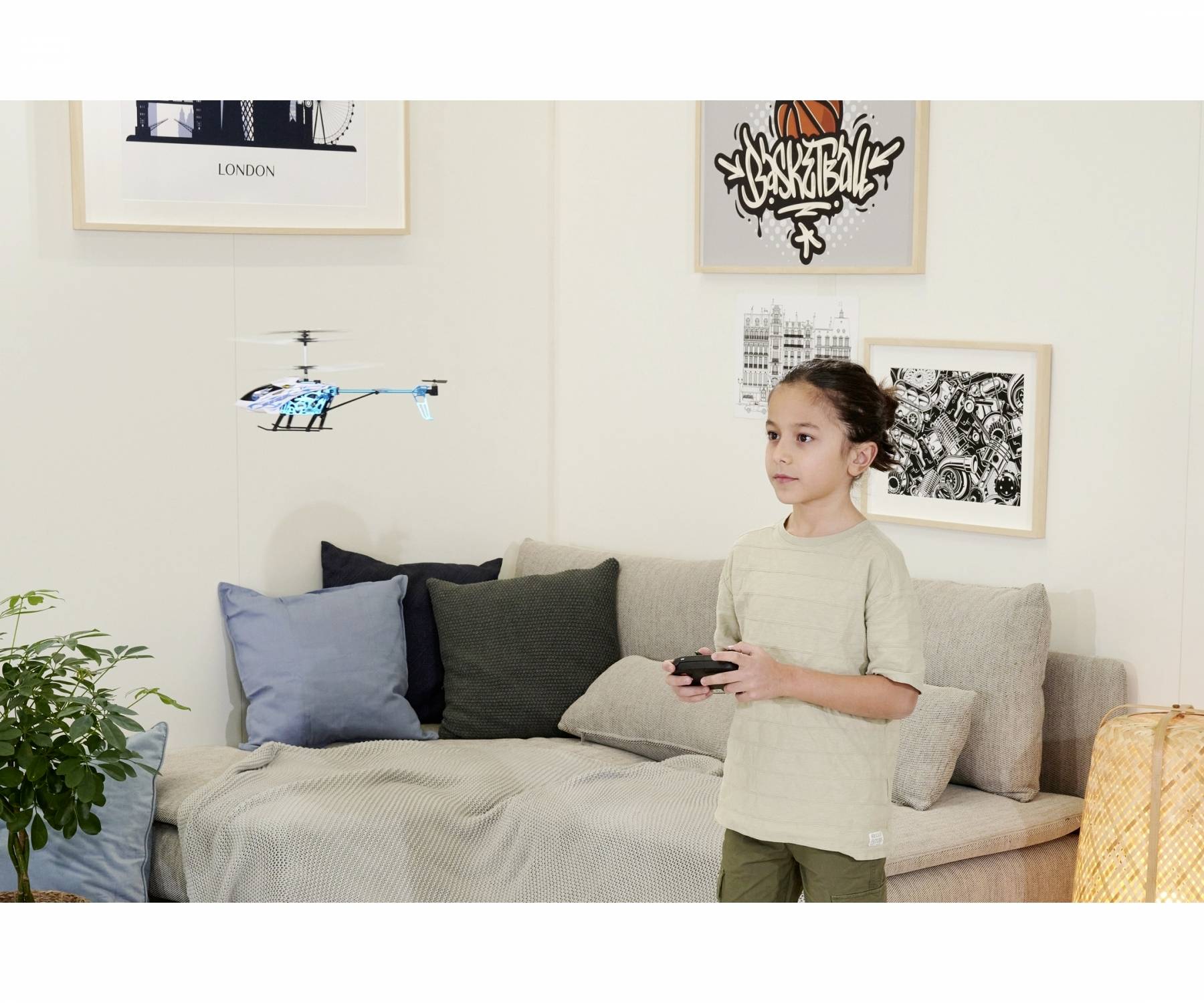 A child is controlling a small remote-controlled helicopter indoors. In the background, pictures are hanging on the wall and a sofa is visible.