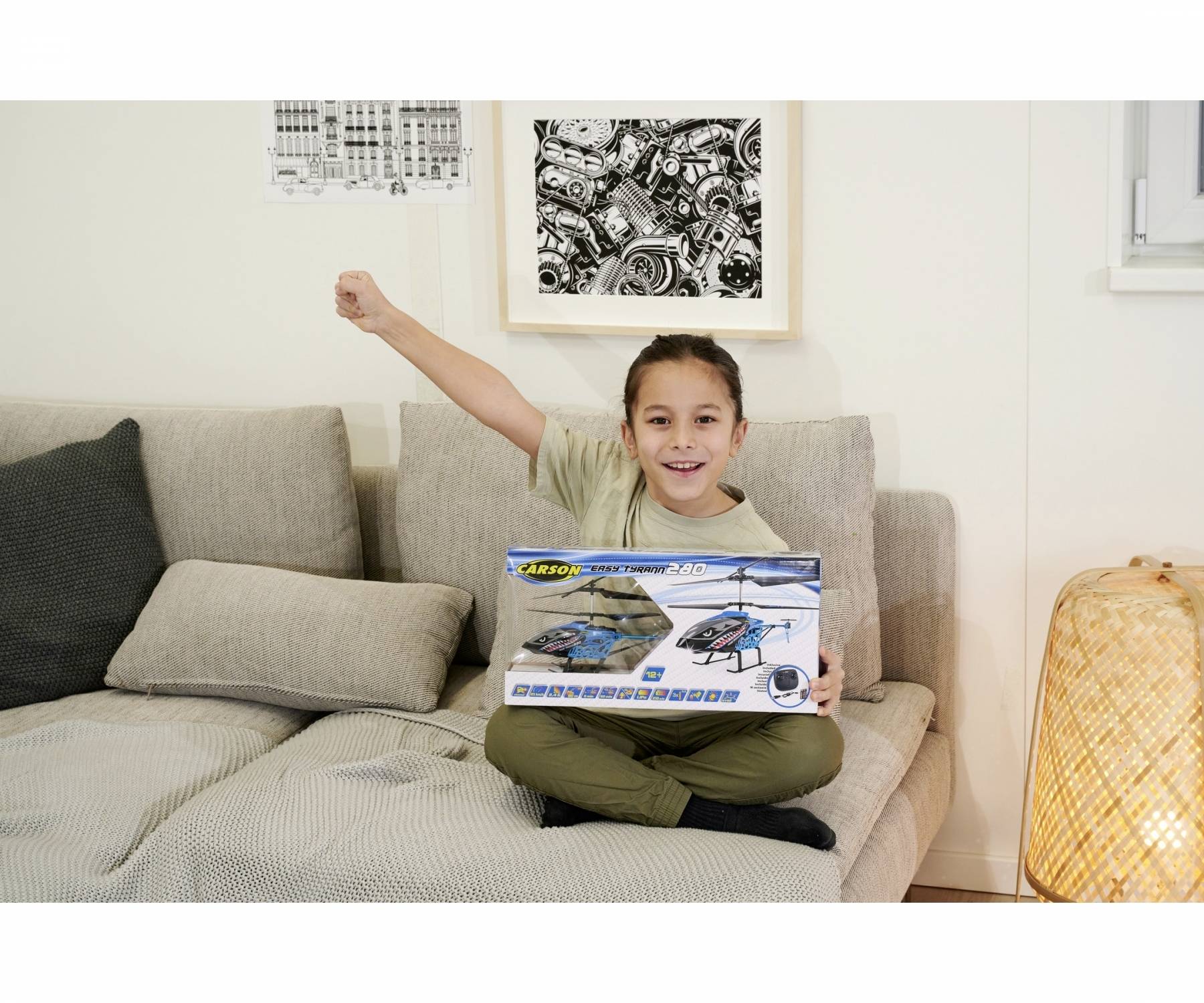 A child sits on a sofa, holding up a toy box with a remote-controlled helicopter with joy. Pictures hang on the wall in the background.