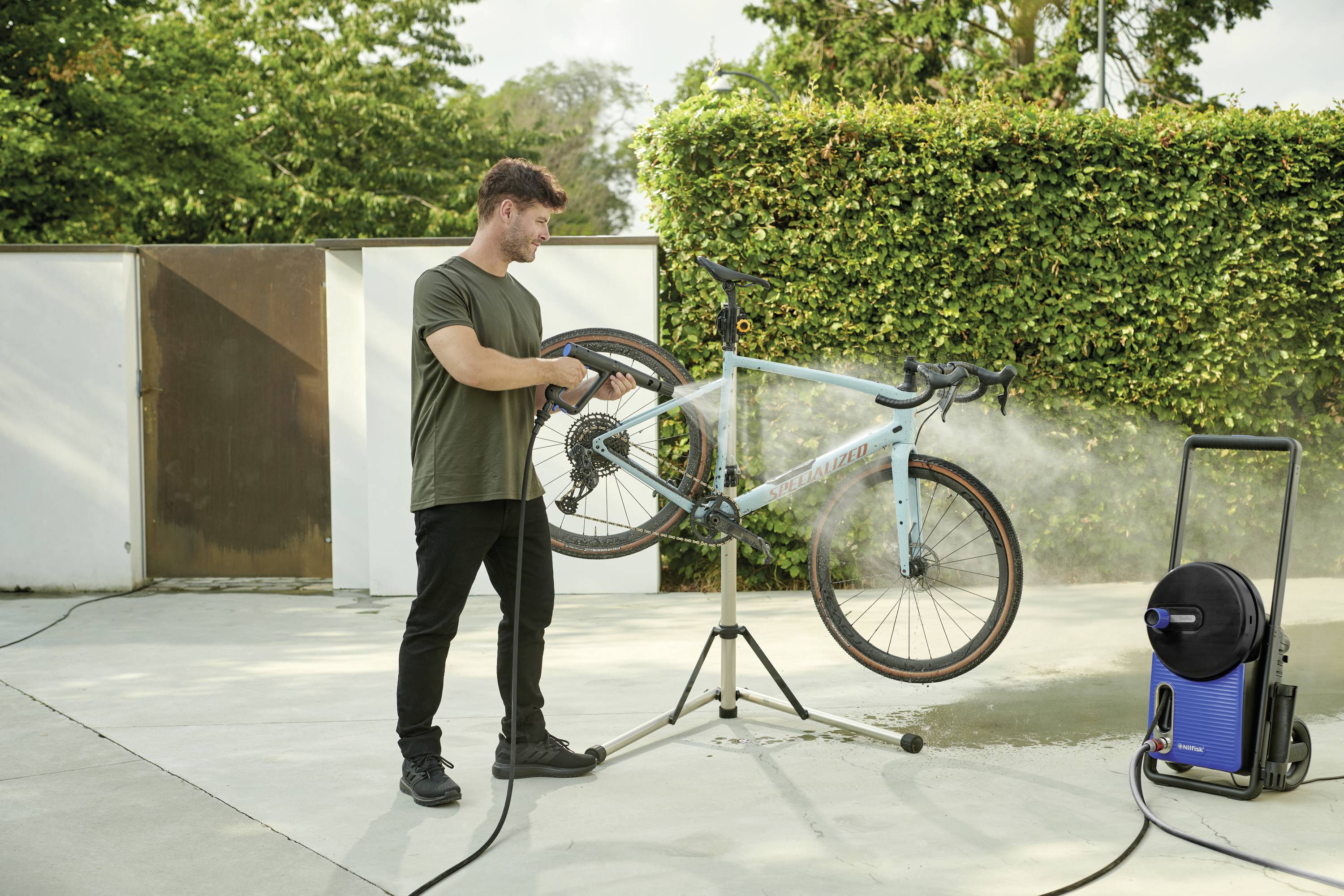 A man is cleaning a bicycle with a pressure washer on a stand outdoors next to a hedge.