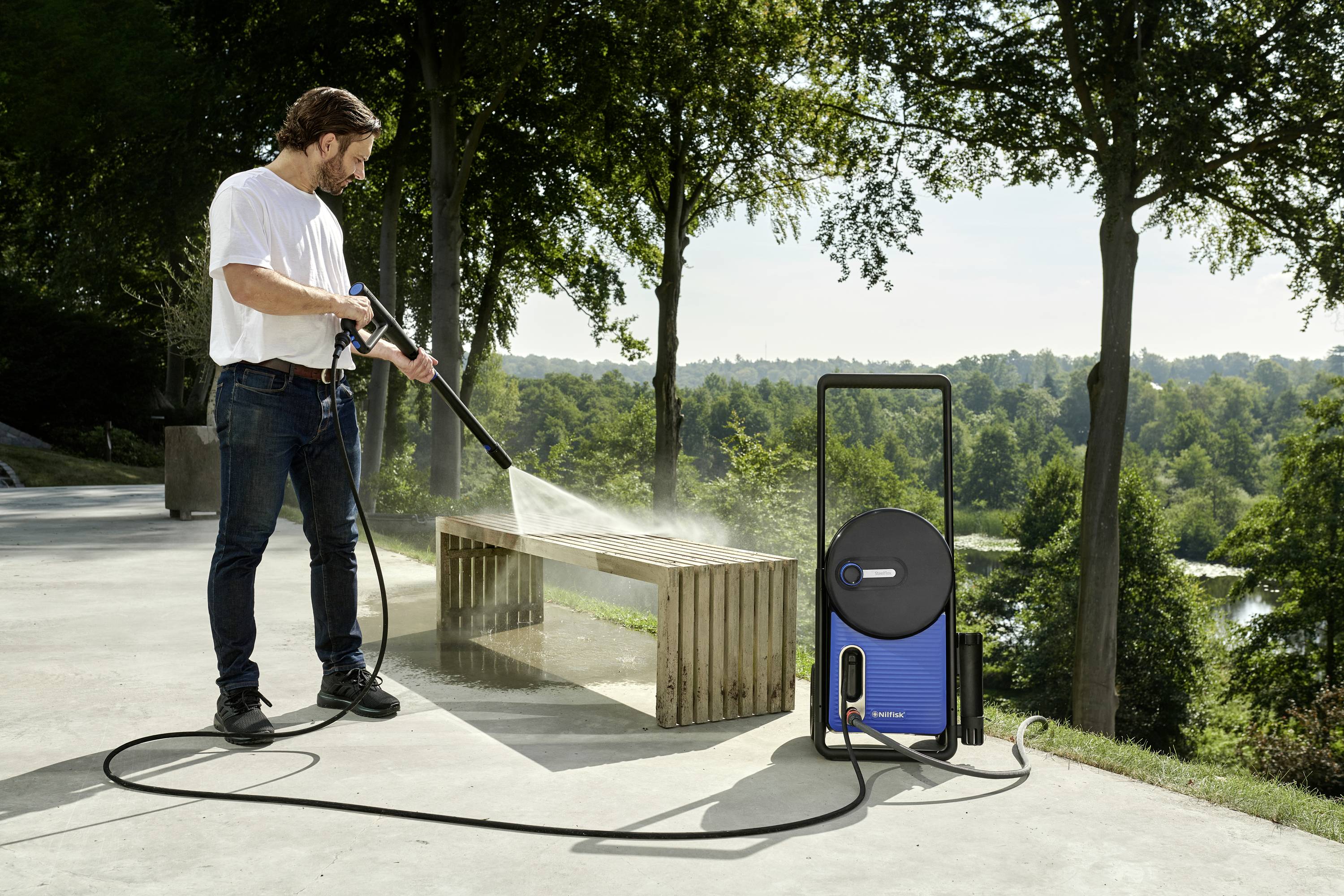 A man in a park is cleaning a wooden bench with a pressure washer. Trees and a green landscape are visible in the background.