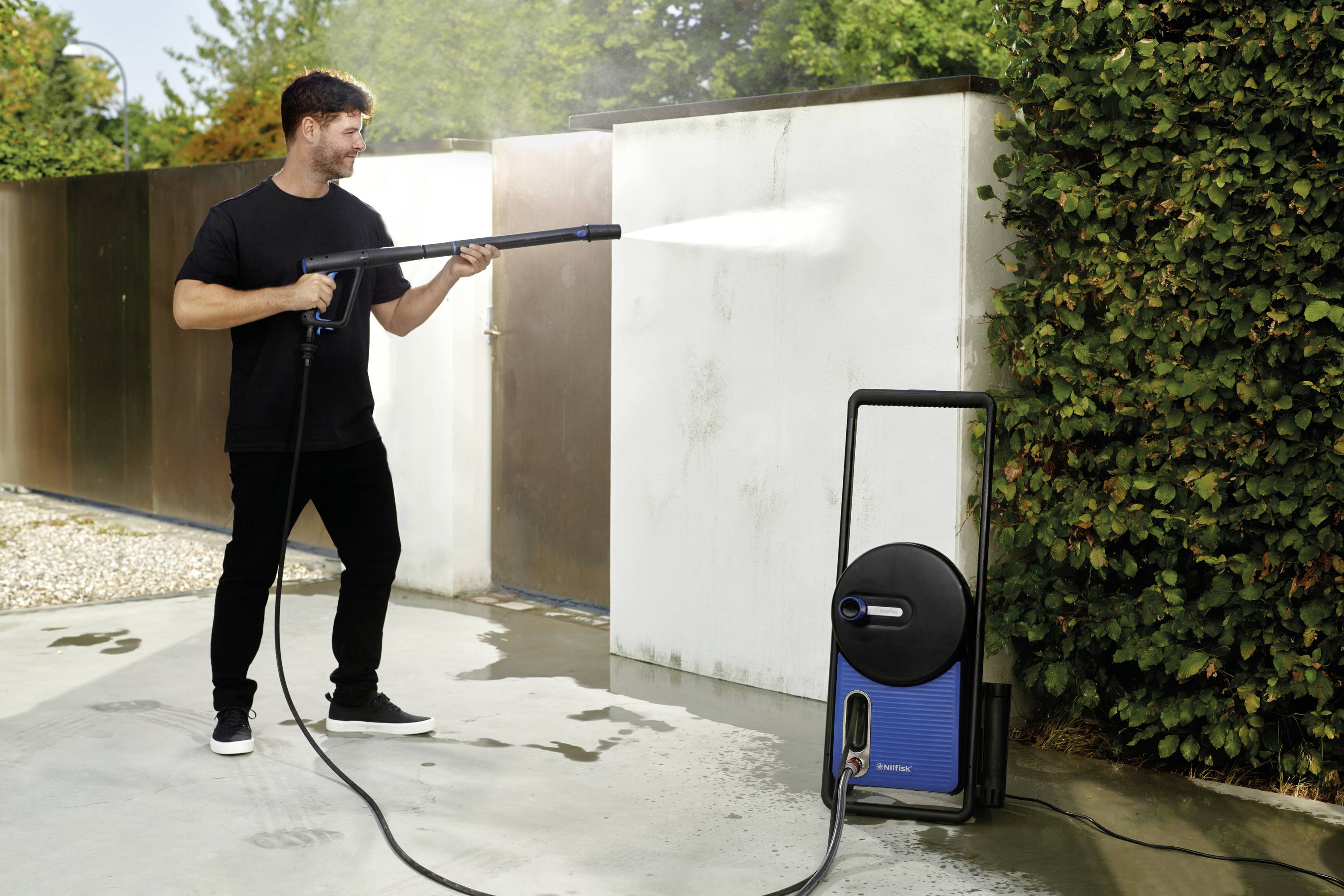 A man is cleaning a white wall with a pressure washer on a terrace. Green bushes and a metal fence are visible in the background.