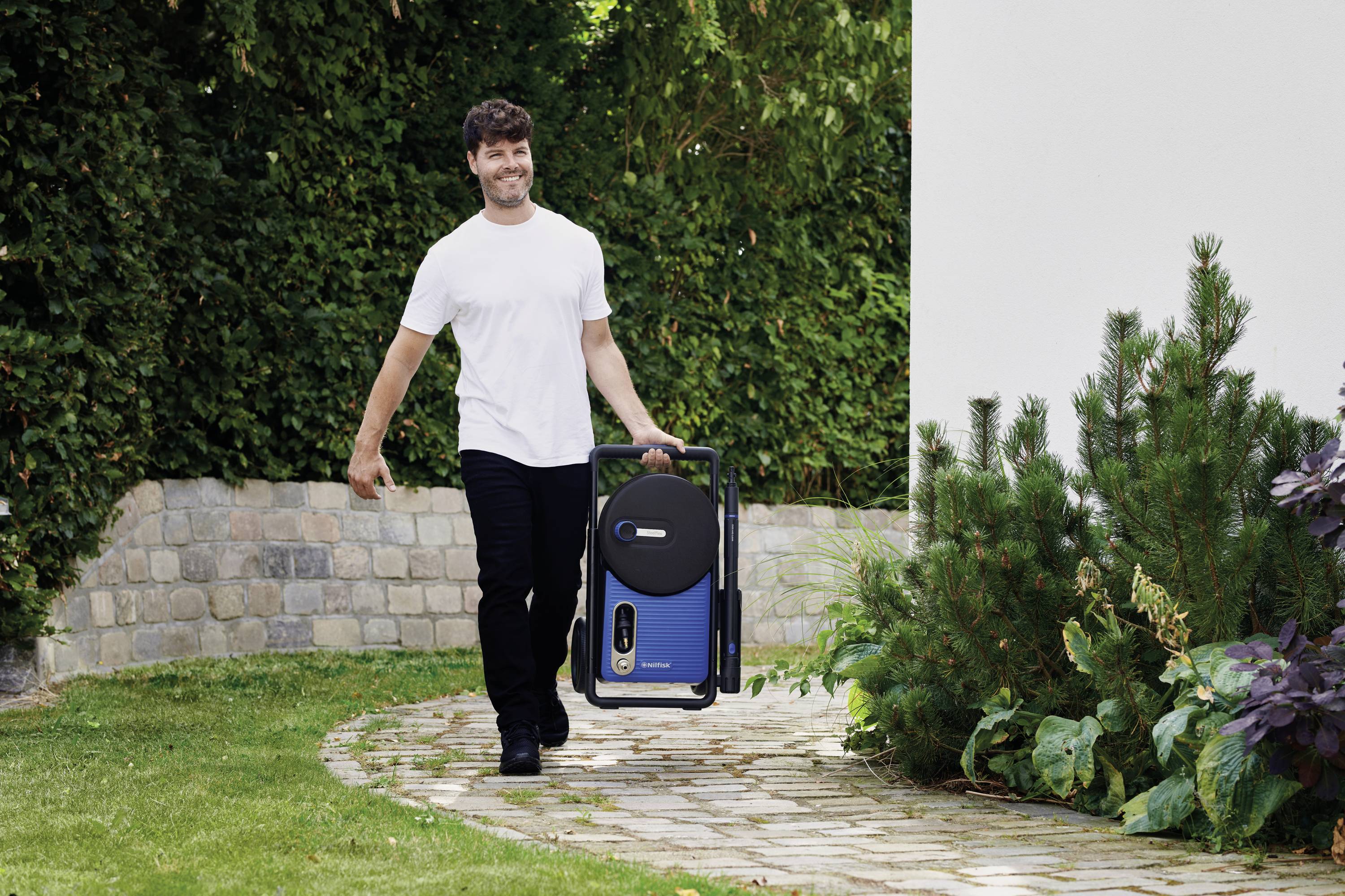 A person is carrying a blue portable charging station along a paved path in a garden, surrounded by plants and a wall.