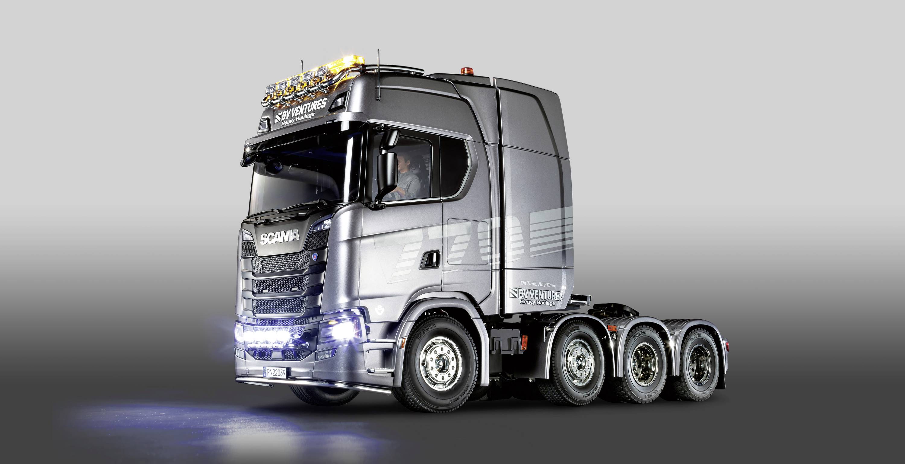 Silver articulated lorry with four axles, illuminated at the front with blue lights. Driver seated in the cab. Company logo on the front.