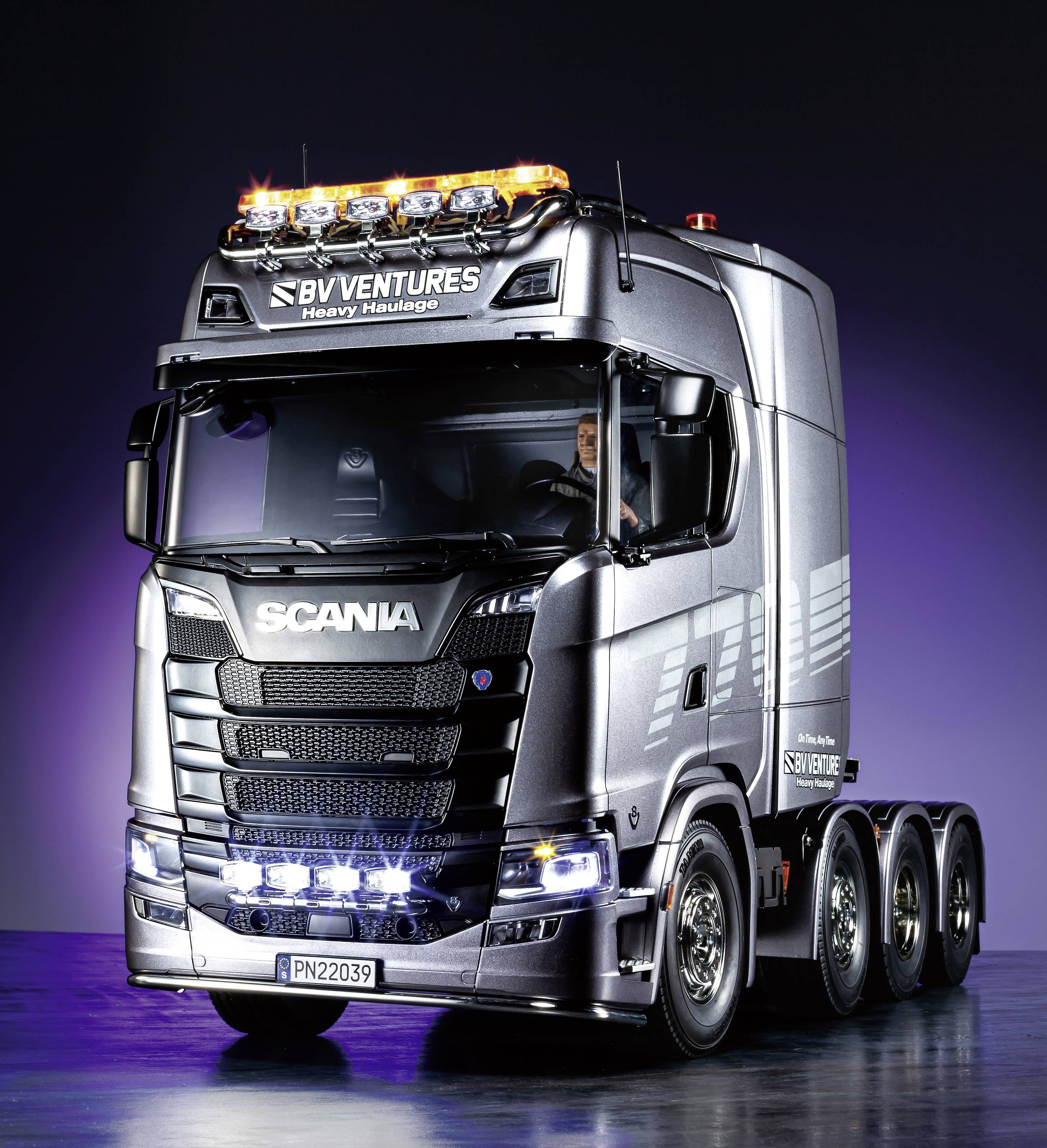 A large, silver-coloured lorry with six wheels and gleaming headlamps against a dark background. The driver is visible.