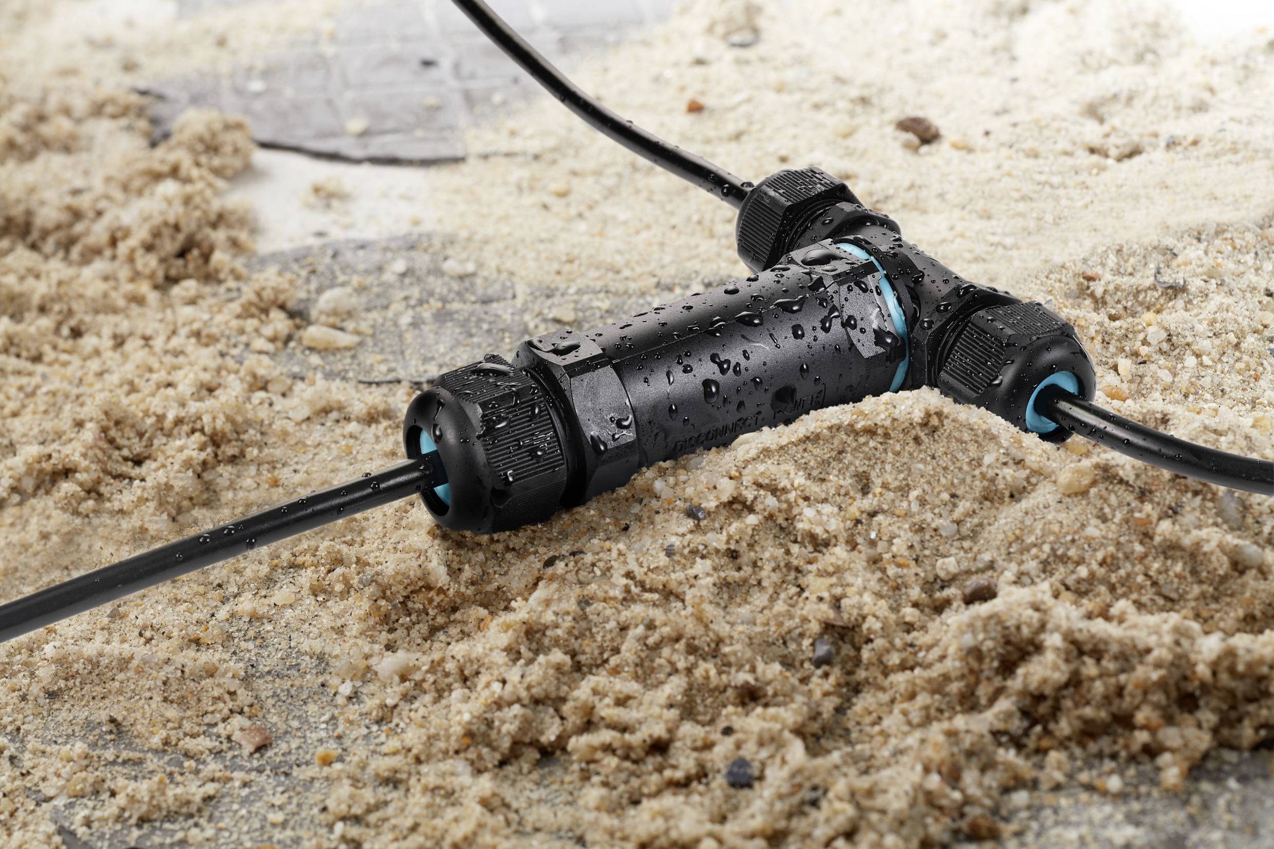 A waterproof cable connector lies on sand and is connected to cables. It demonstrates that it is suitable for outdoor use.