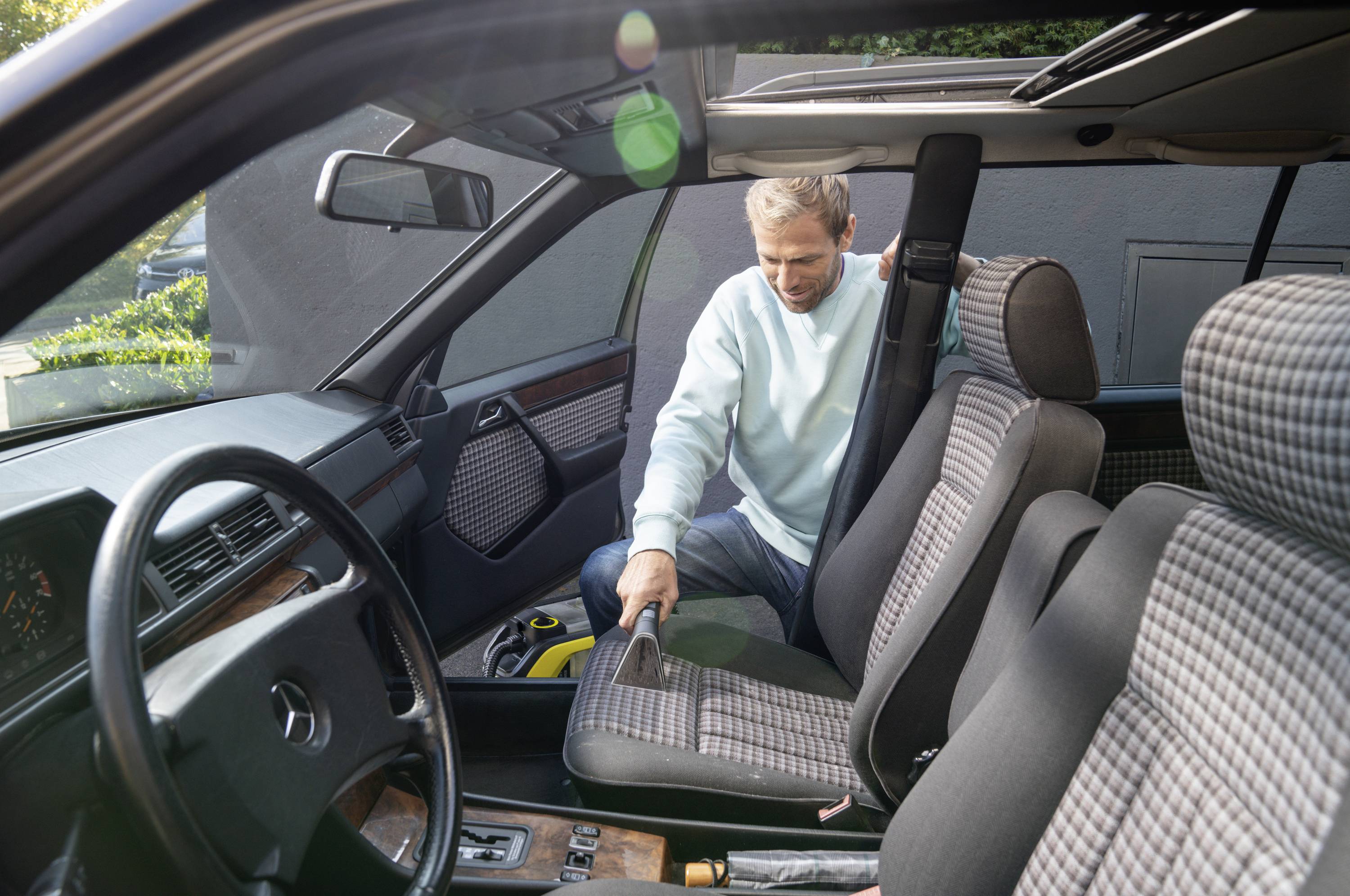 A man is cleaning the interior of a car, vacuuming the floor. He is wearing a light blue jumper. Daylight is coming through the car window.