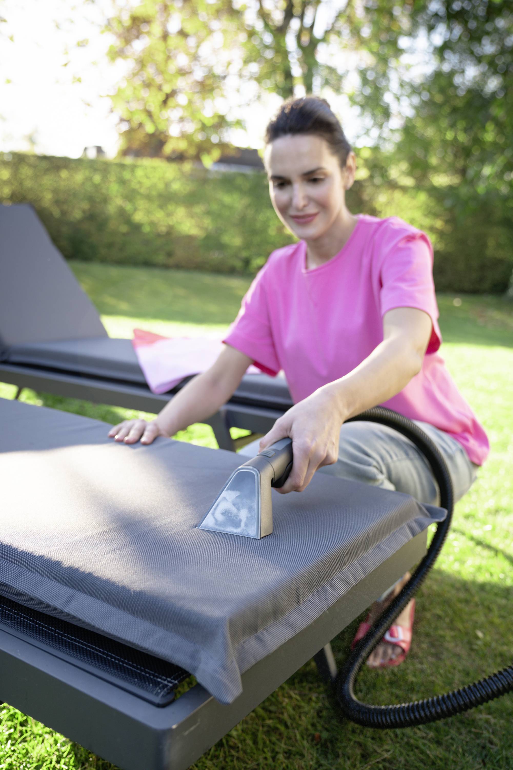A woman is cleaning the cushion of a sun lounger with a steam cleaner in the garden. Green vegetation can be seen in the background.