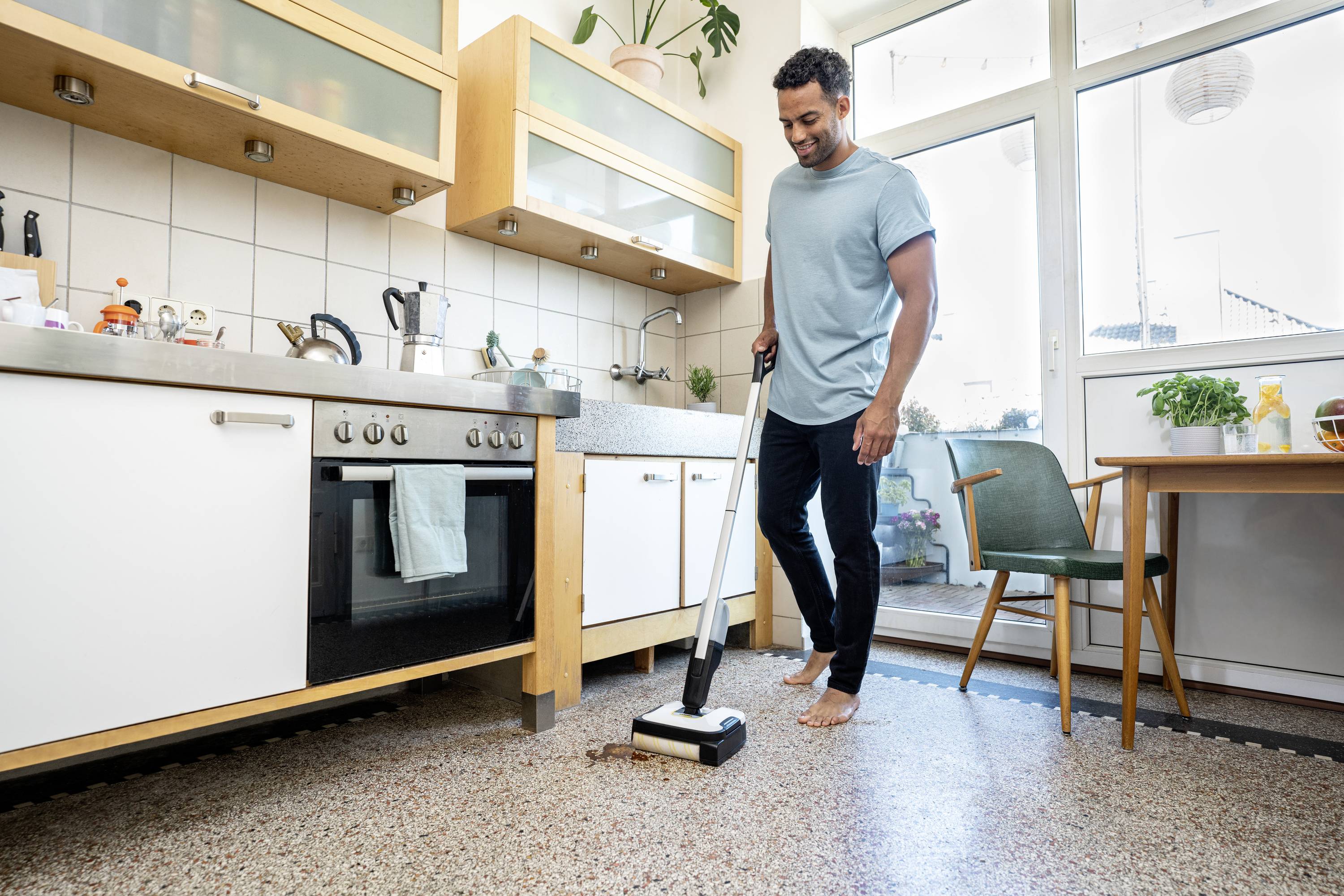 A person is cleaning a kitchen floor with a mop. The kitchen is modern, furnished with wooden furniture and large windows.