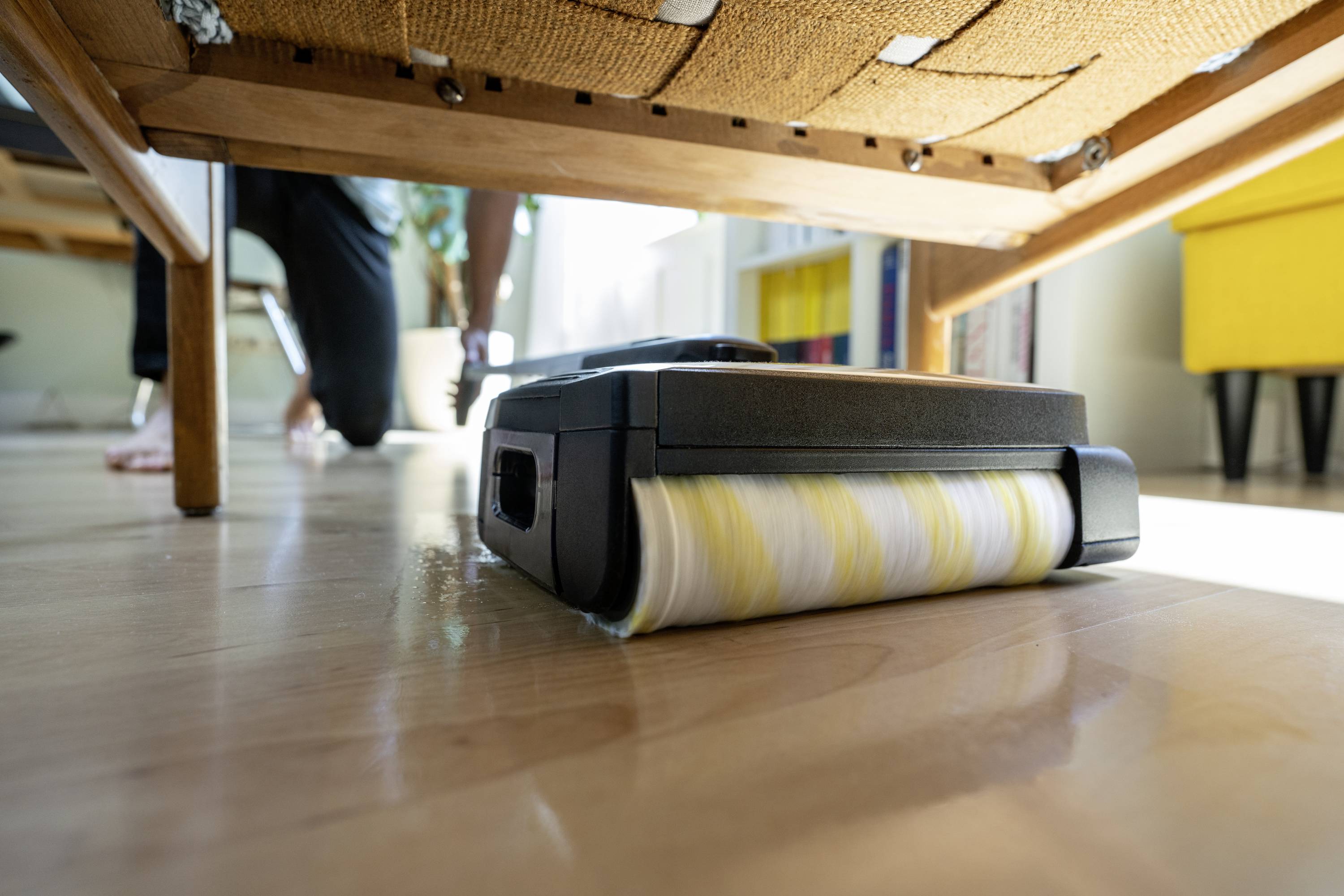 A robot vacuum cleaner is cleaning the wooden floor underneath a sofa. A person is blurrily visible in the background.