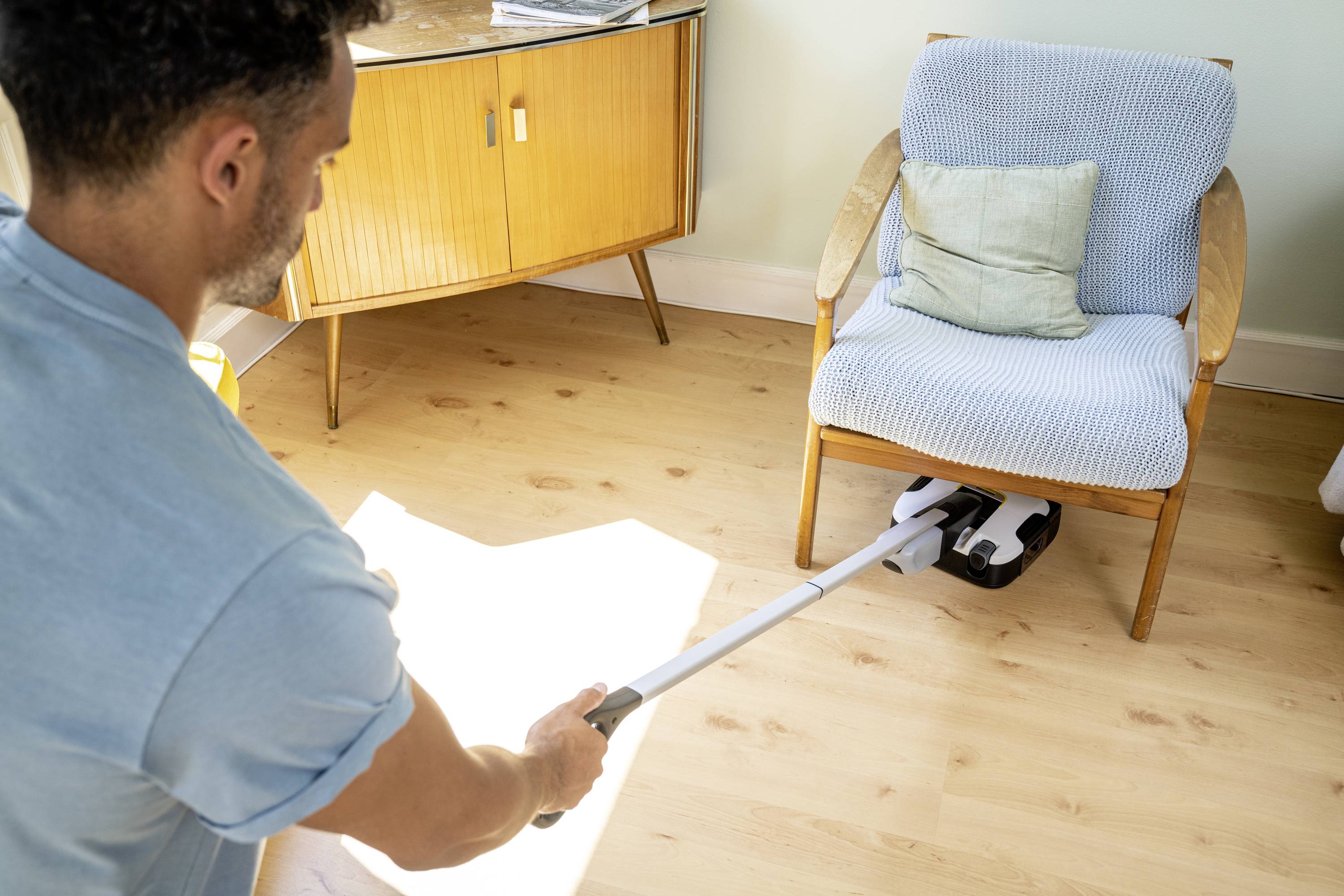 A man is vacuuming a wooden floor beneath a chair in a bright room. A sideboard stands in the background.