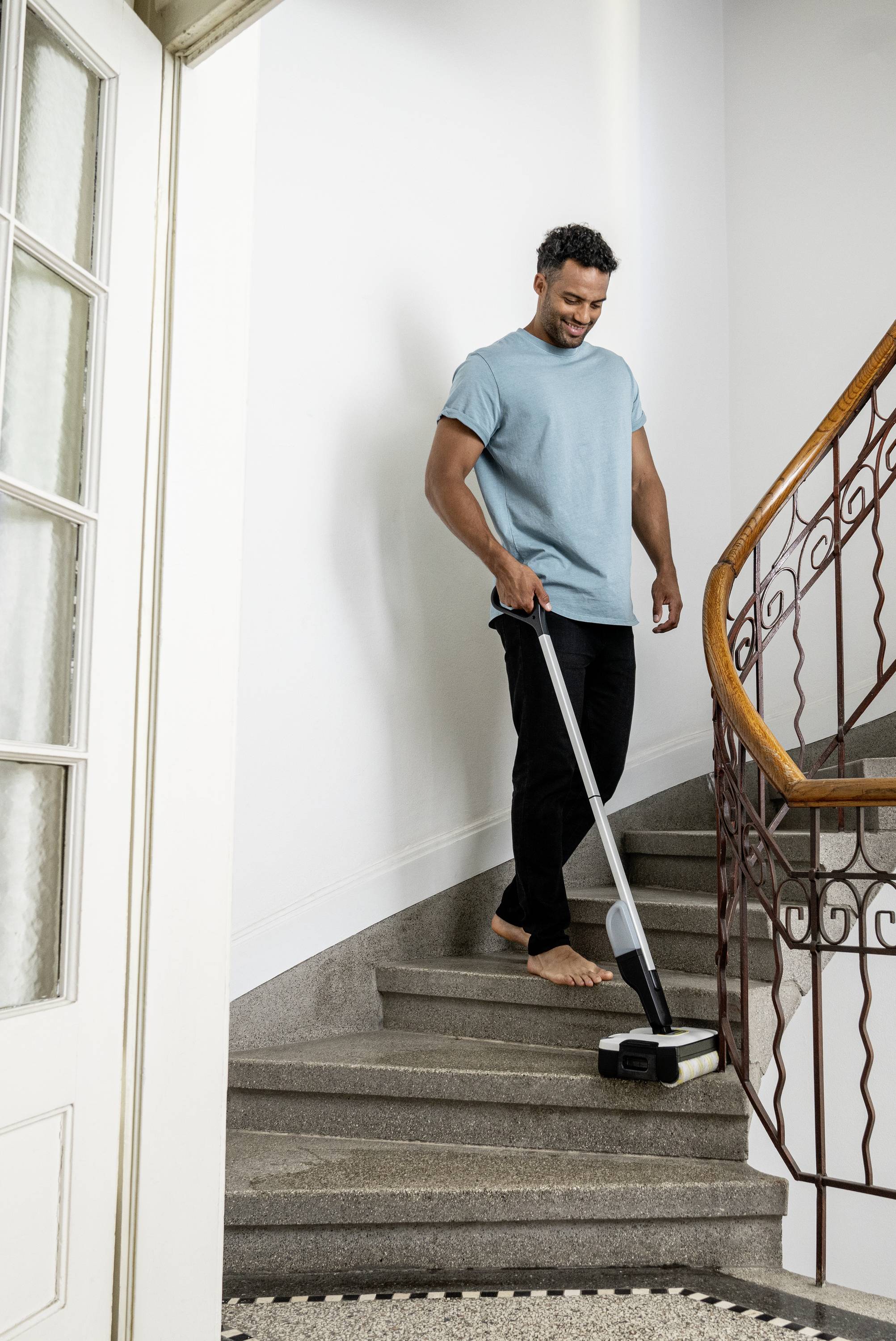 A person is hoovering barefoot up the stairs of a residential house. They are wearing a light blue T-shirt and black trousers.