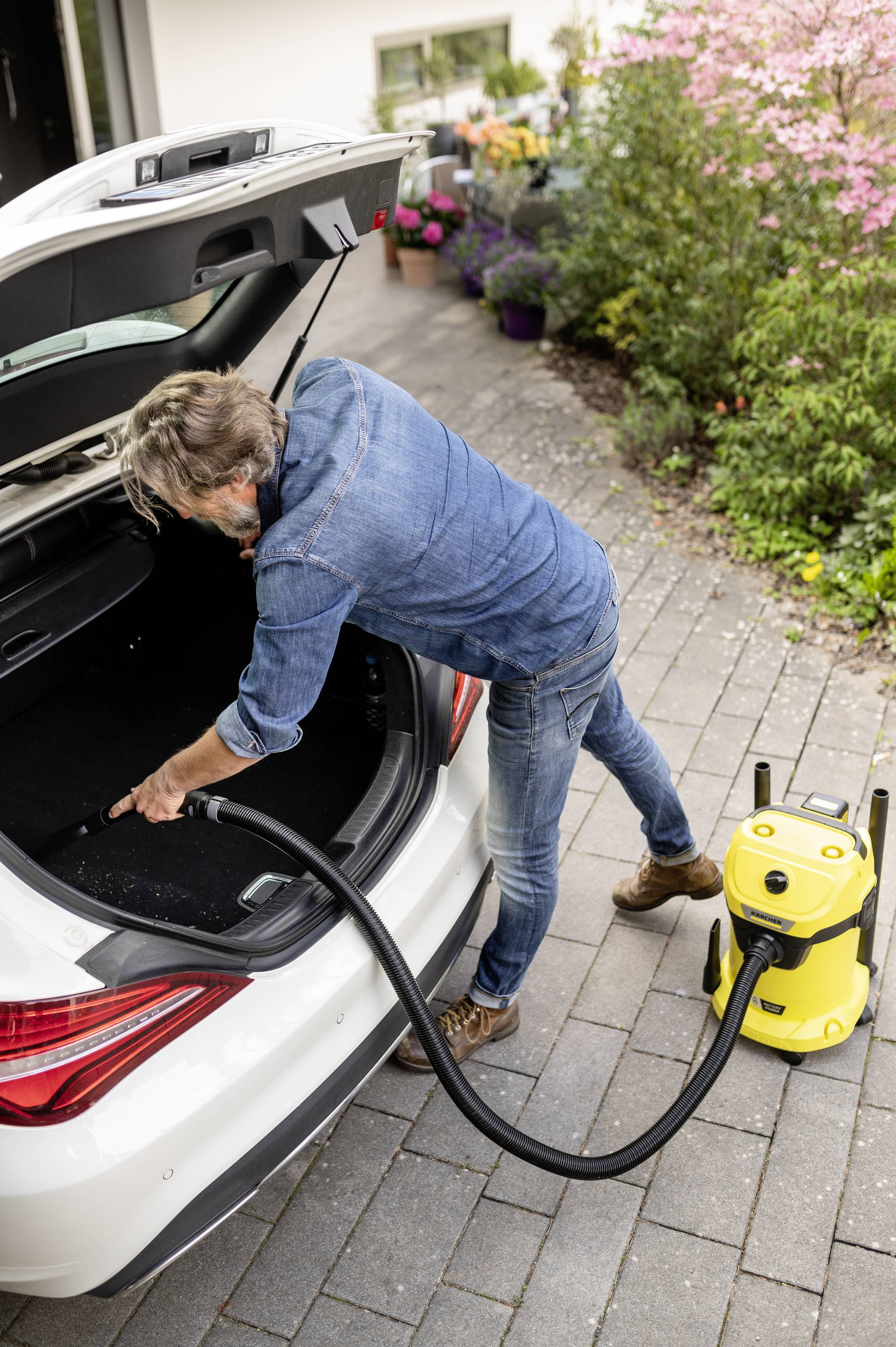 A man is cleaning the boot of a car with a yellow hoover. Plants and flowers can be seen in the background.