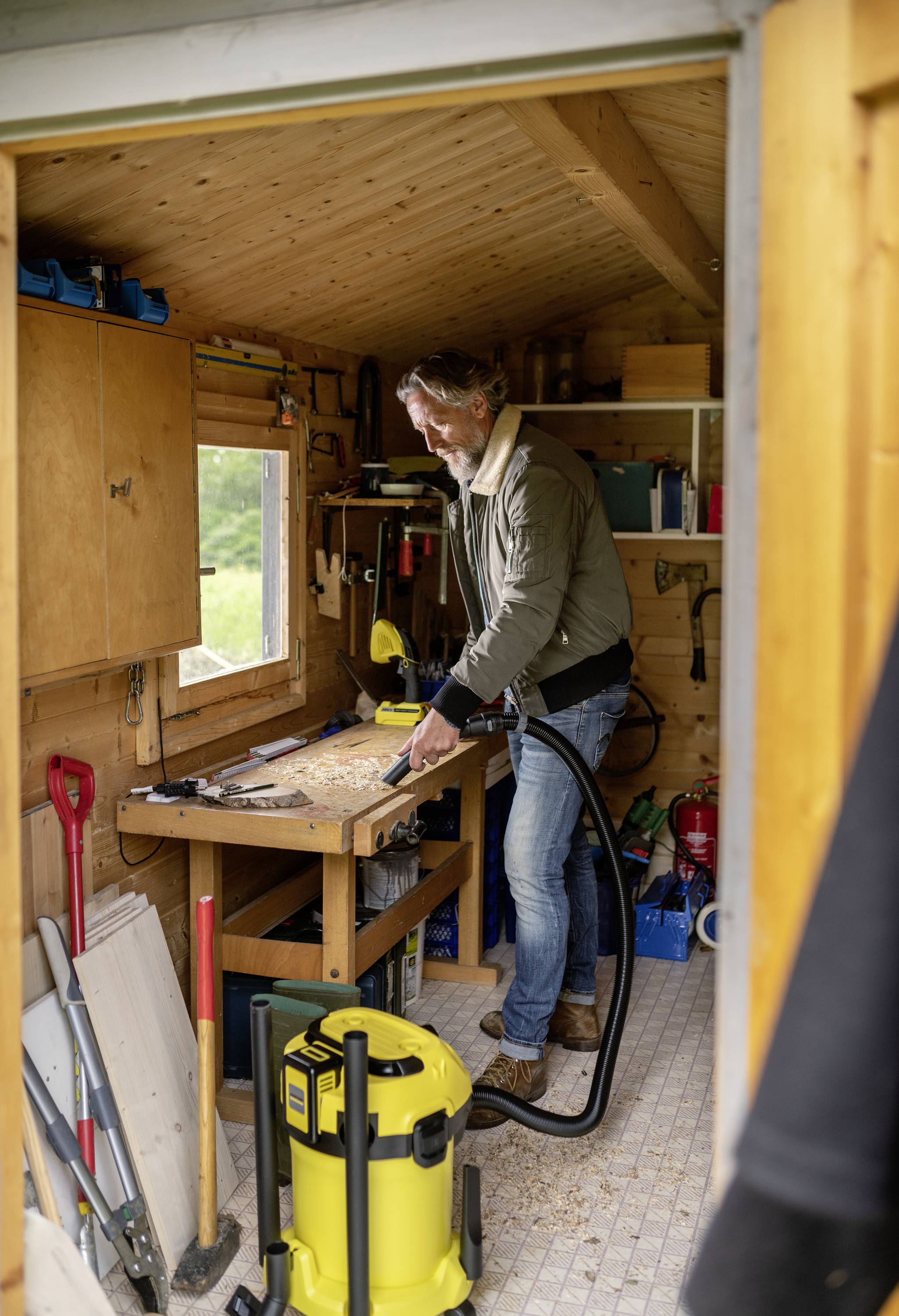 A man is working in a woodwork workshop. He is standing at a workbench and sawing a piece of wood. Tools and equipment are scattered around him.