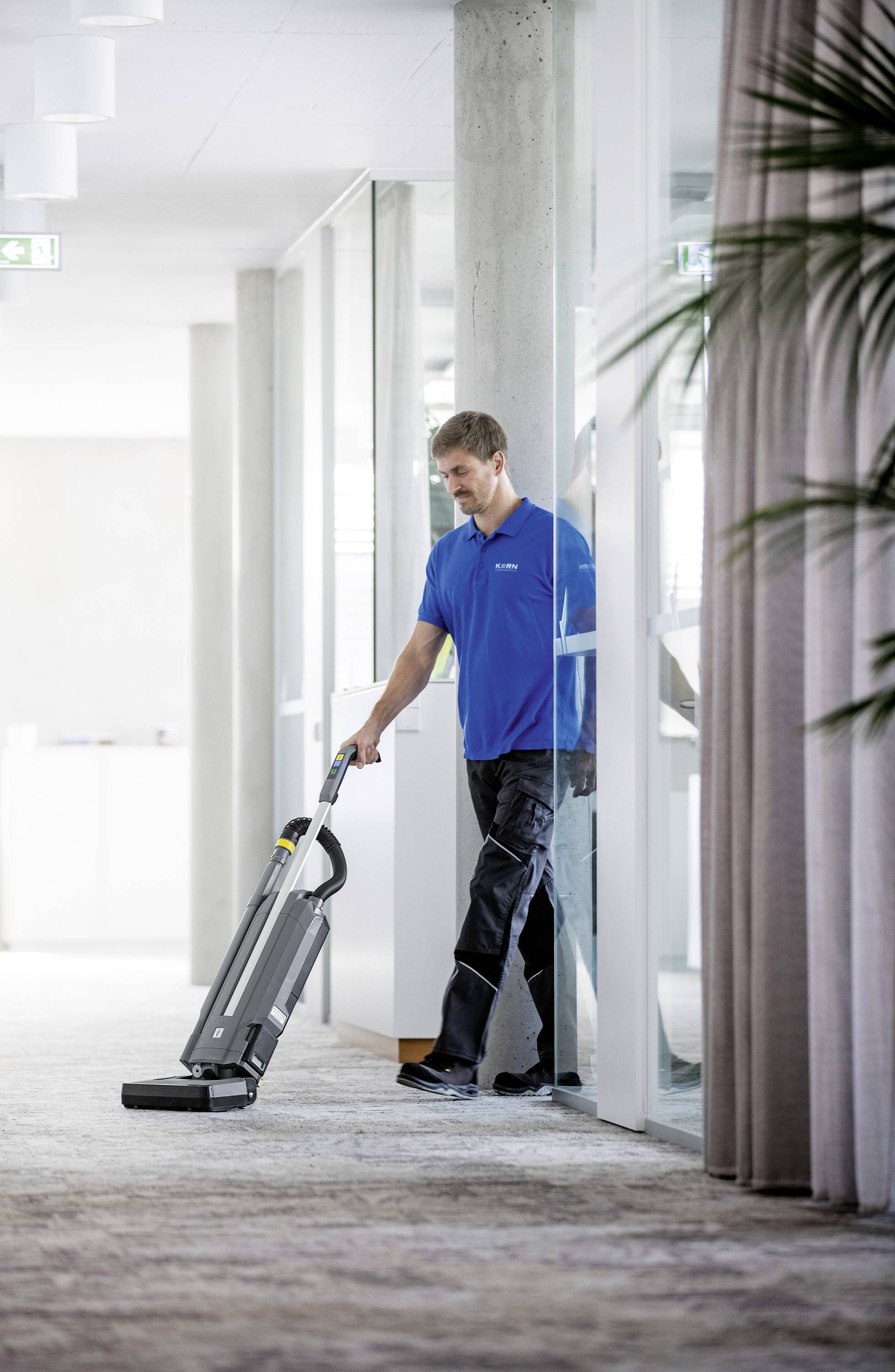 A person is hoovering the carpet in a modern office building with glass walls and plants in the foreground.