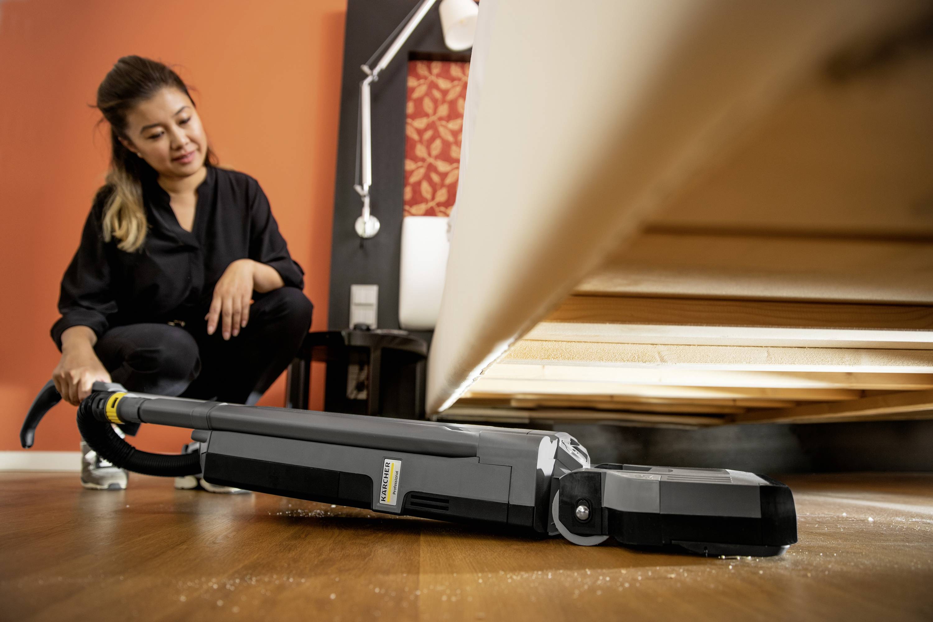 A person is vacuuming underneath a sofa to remove dust. The vacuum cleaner is flat enough to reach the area.