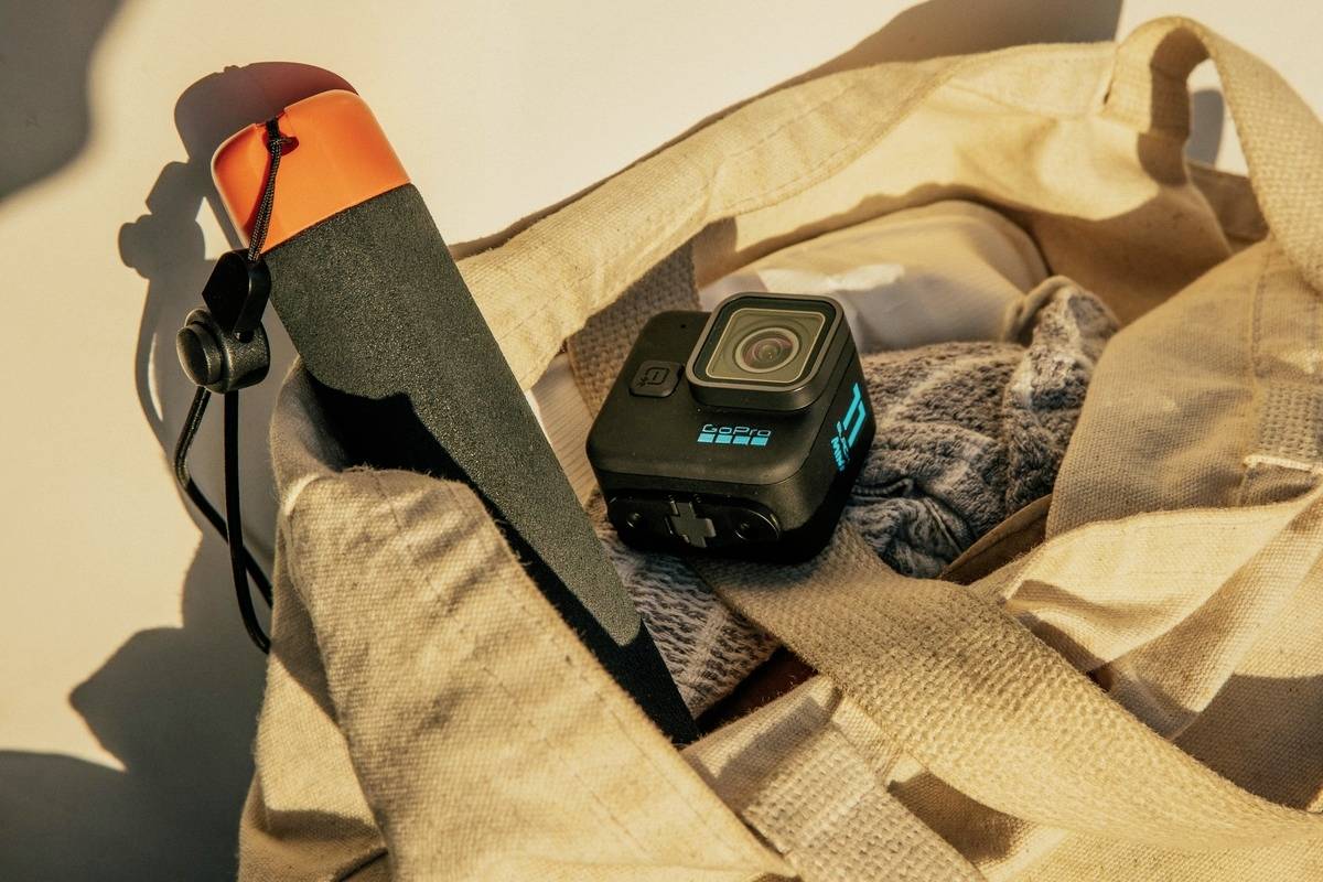 A beige fabric bag contains a camera and an orange-black handheld device. The bag is resting in warm sunlight.