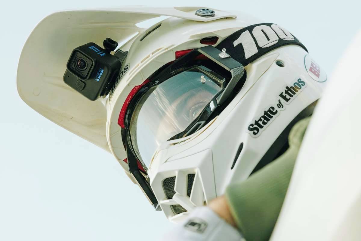 A motorcyclist wearing a helmet with a mounted camera looks ahead intently. The helmet bears the inscription 'State of Ethos'.