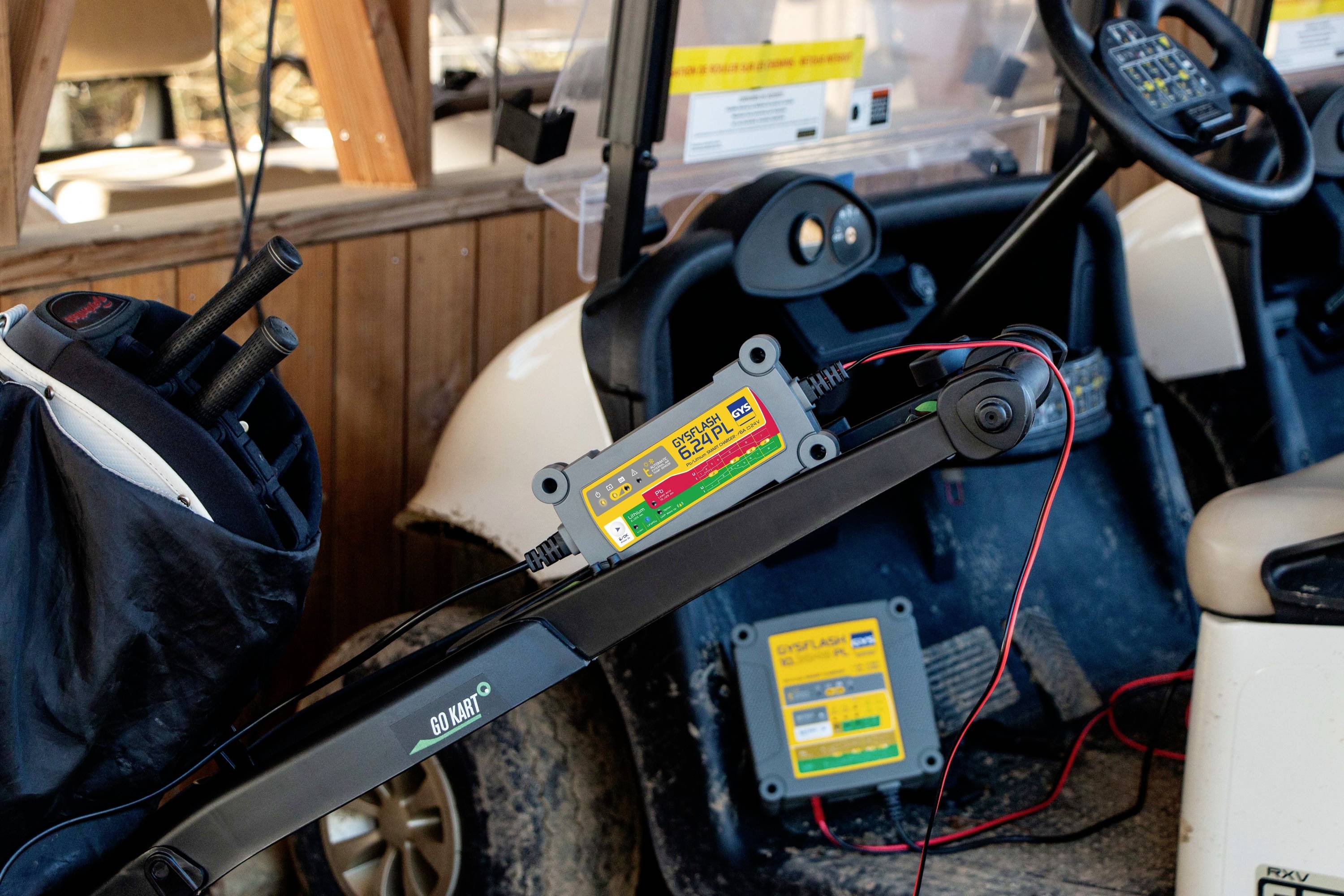 An electric golf buggy is being connected to a charger. Golf bags and clubs are visible in the background.