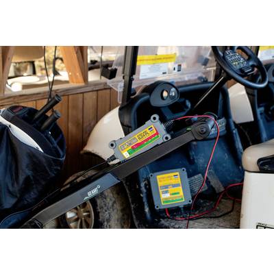 Golf vehicle in a garage, connected to a battery charger. Various equipment is visible in the background.