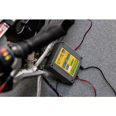 A battery charger is connected to a bicycle standing on a carpet floor. The display shows charging process details.