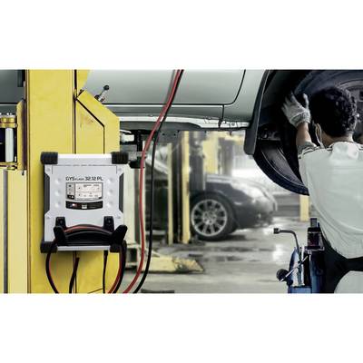 A mechanic is working on a raised car in a workshop. A battery testing device is attached to a support post.