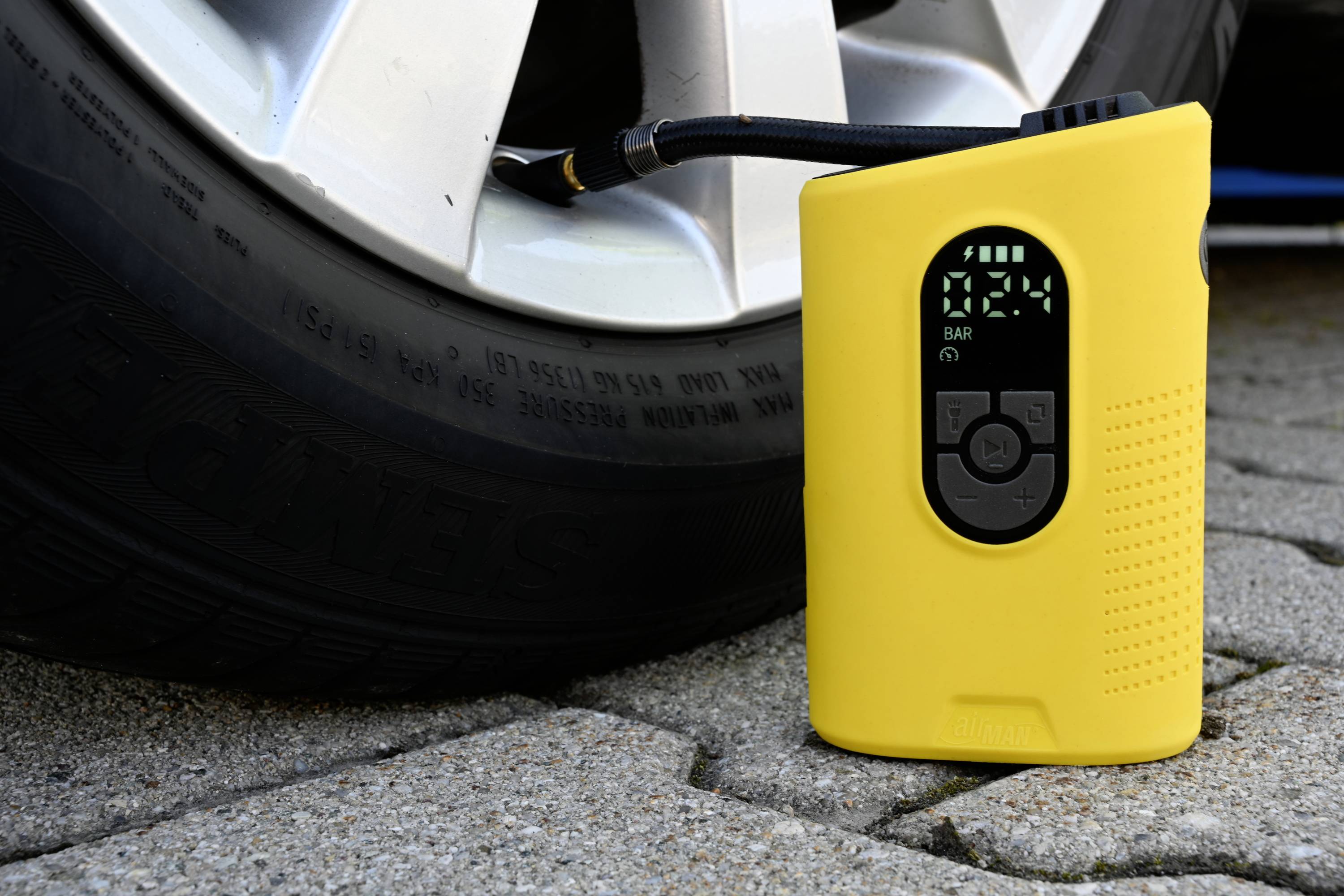 Yellow digital tyre inflator displays tyre pressure in bar, connected to a car tyre on paving stones.