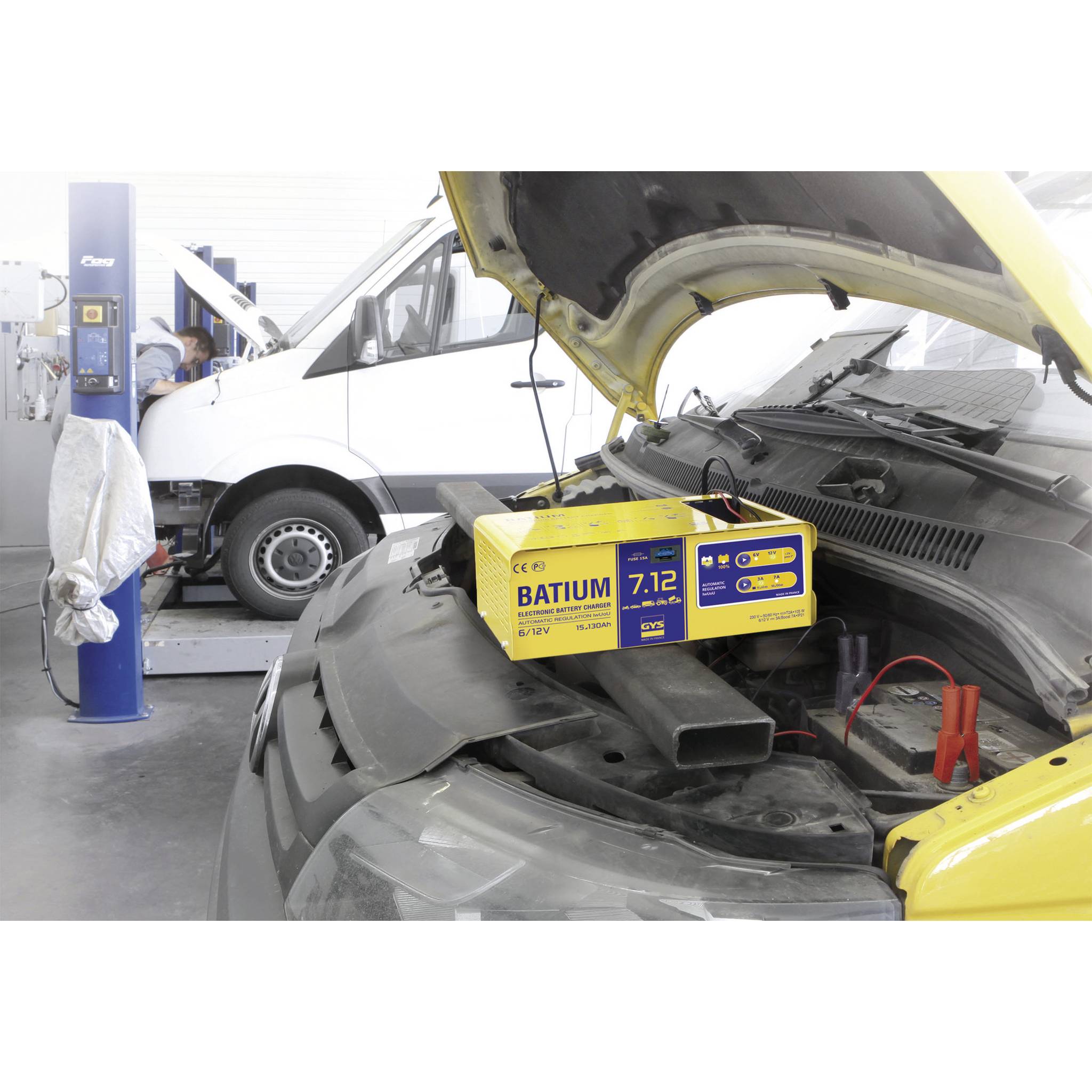 A car in a workshop with its bonnet open, with a yellow BATIUM battery charger connected to it.