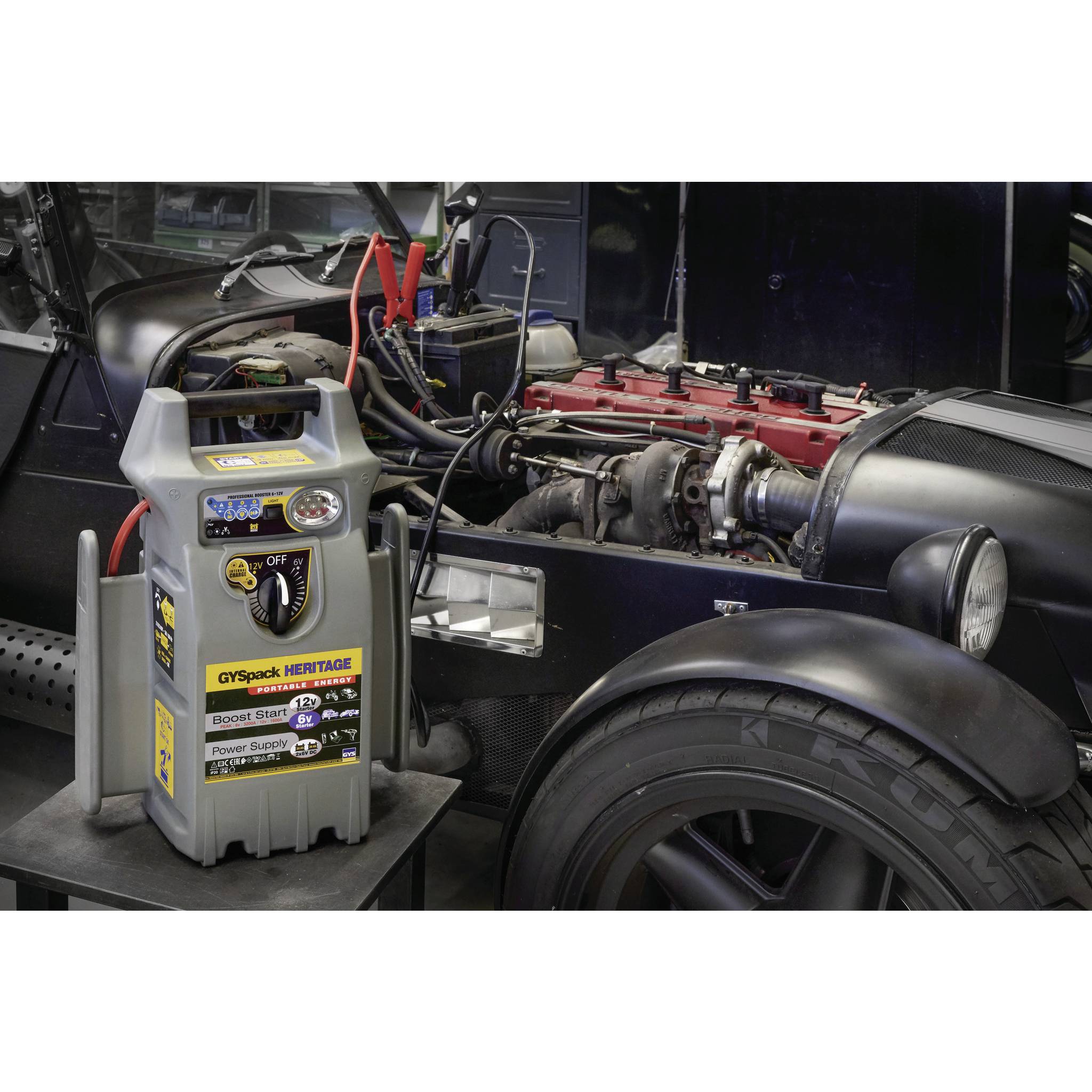 A car parts and battery booster is positioned next to an open engine bay of a classic car, connected for power supply.
