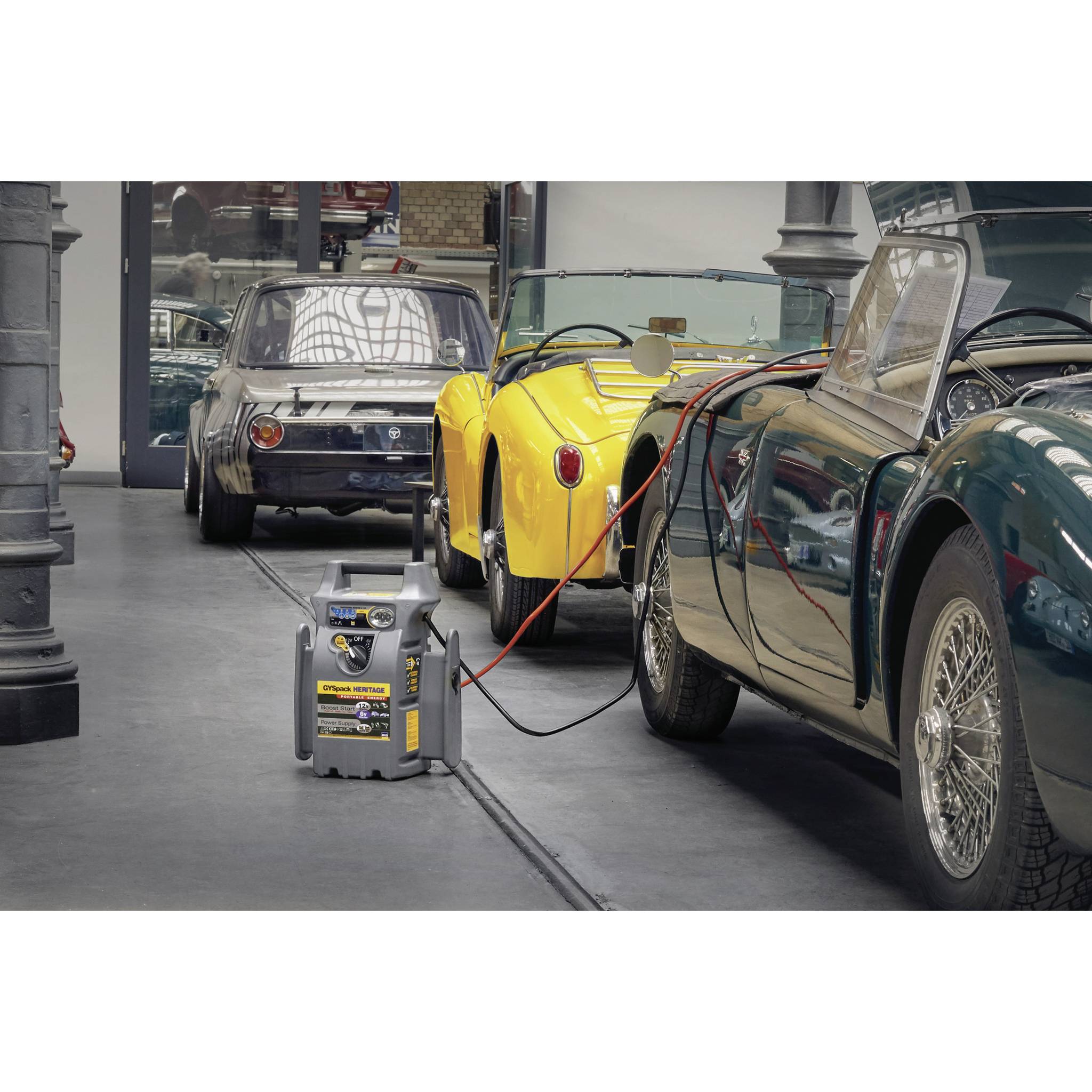 Classic cars are parked in a workshop, connected to battery chargers. In the foreground, a battery charger sits on the floor.