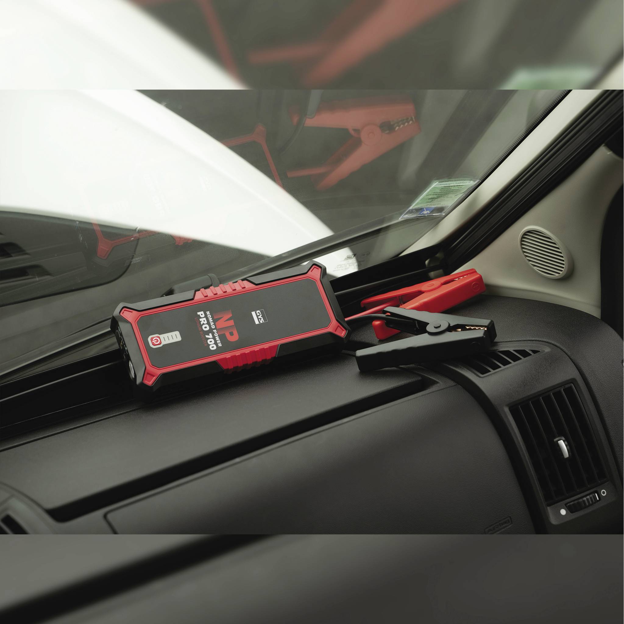 A portable jump starter is lying on the dashboard of a car. Jump leads are connected and ready for use.