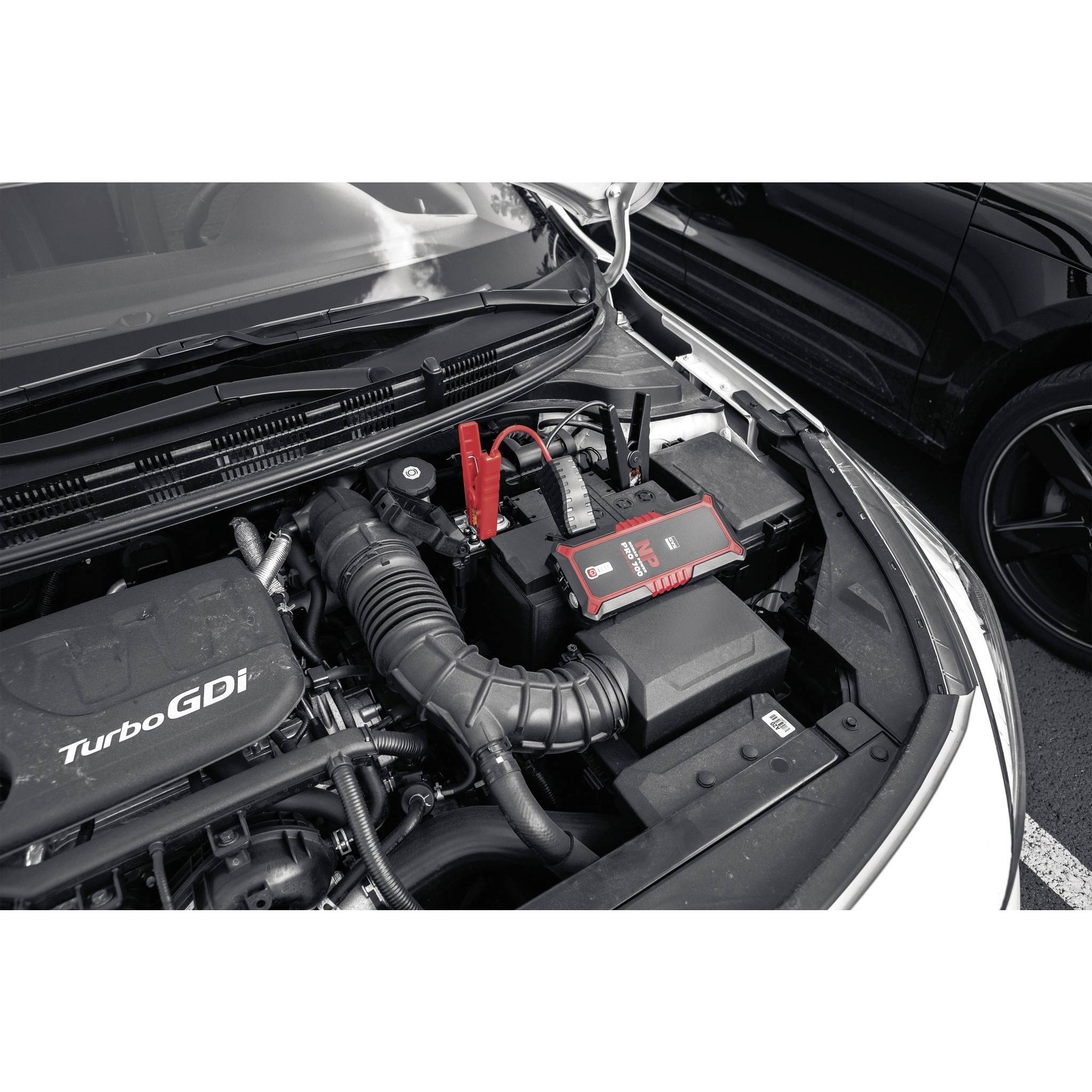 A car boot with an open bonnet, showing a car battery with a jump starter and cables connected.