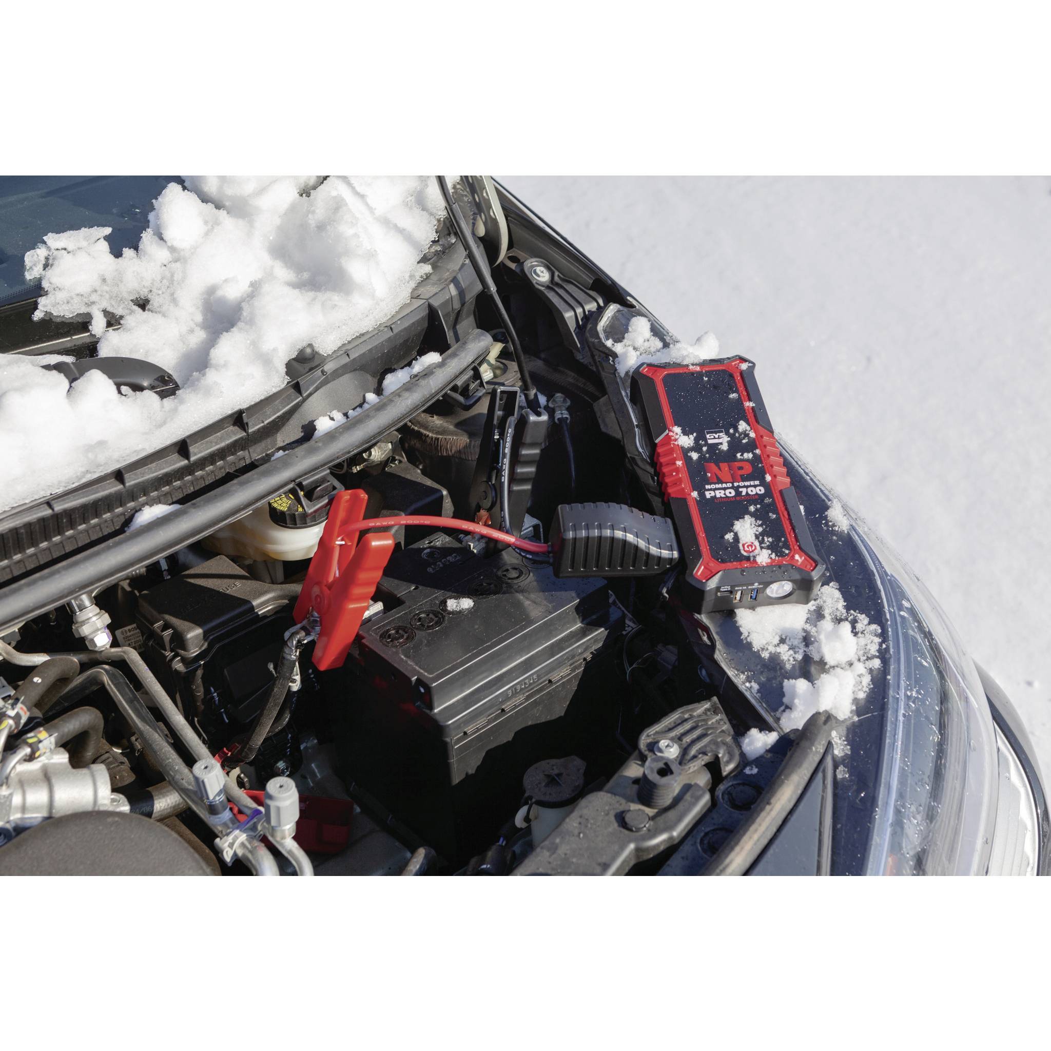 A car in the snow with a red jump starter battery connected to the car battery. Main purpose: Starting the car in cold weather.