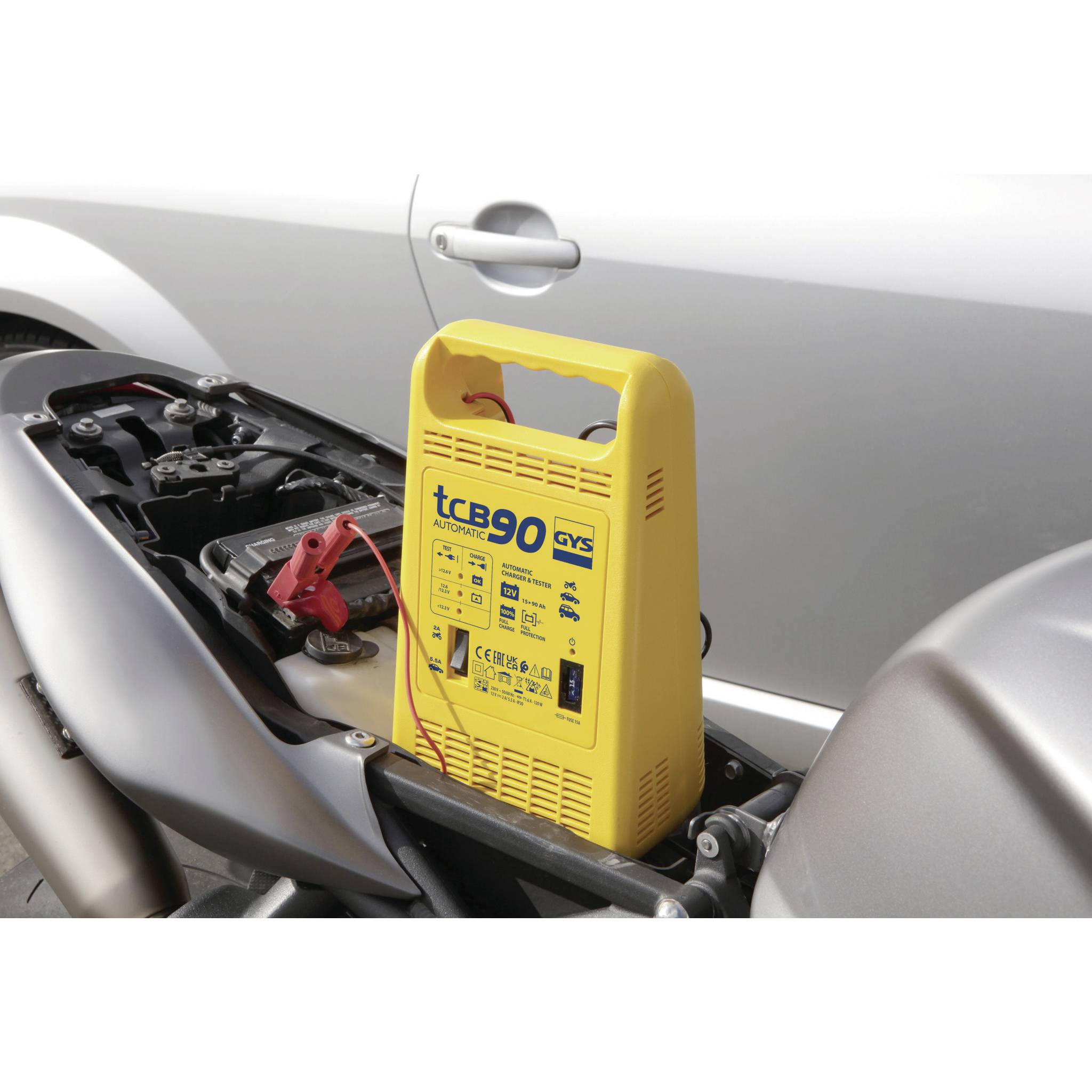 A yellow battery charger is positioned next to the open motorcycle battery. Cables are connected to the battery terminals.