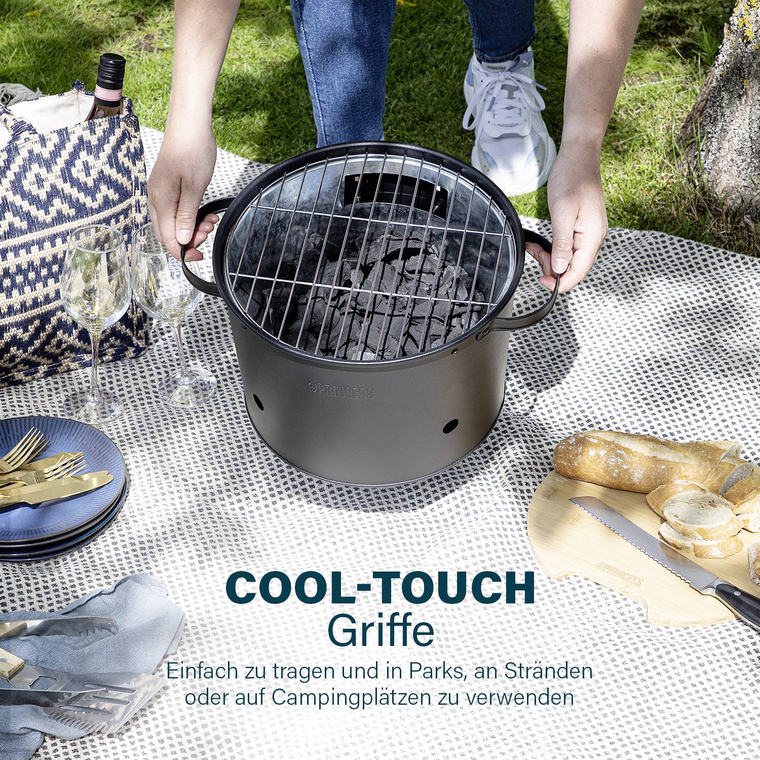 A person is positioning a portable barbecue on a picnic blanket outdoors. Beside it are cutlery, bread, and a wine basket.