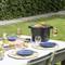A table in the garden with laid-out crockery, glasses, and plates. A barbecue in the background with food on it. Sunny day.