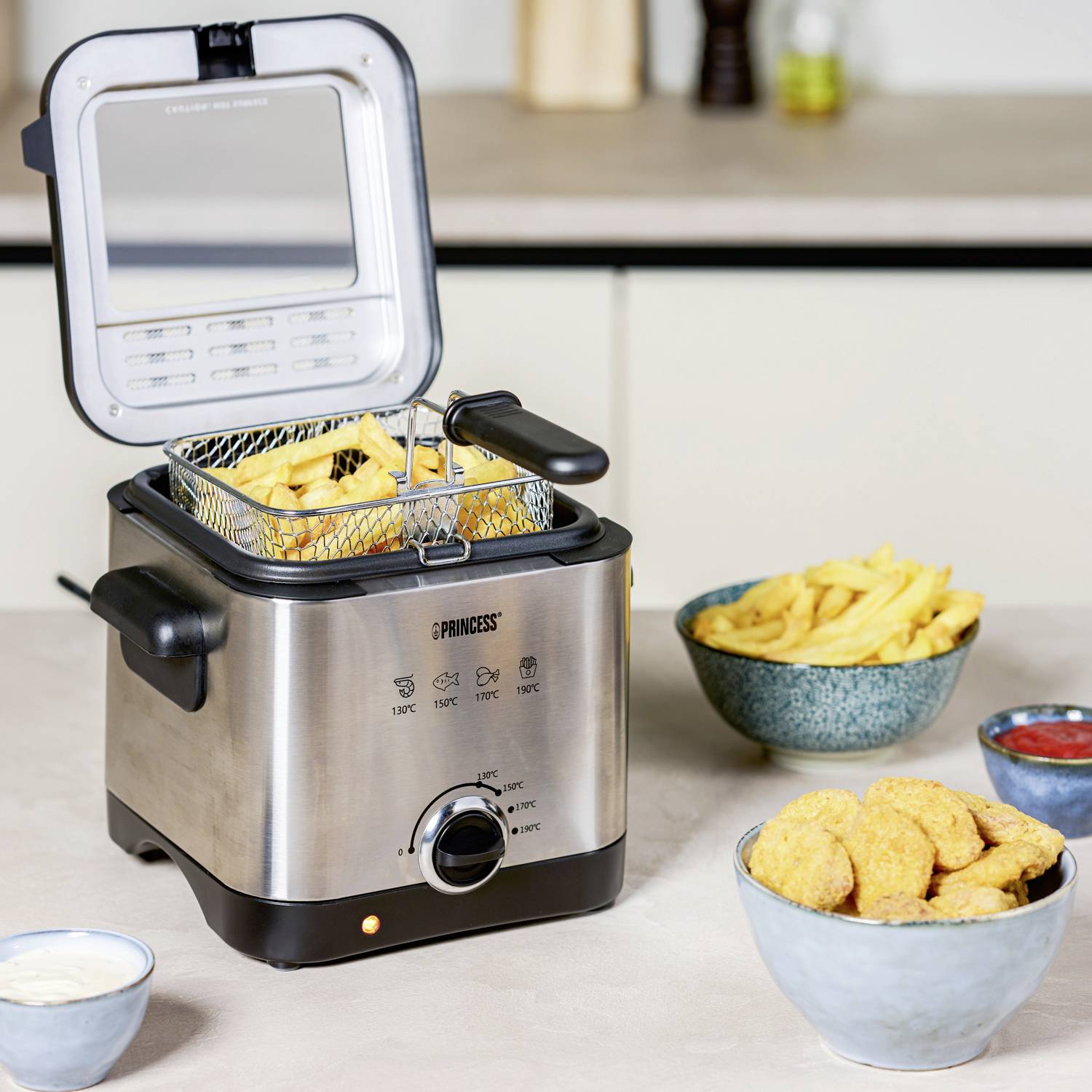 A deep fat fryer with its lid open, filled with chips. Beside it are dishes of chips and chicken nuggets on the worktop.