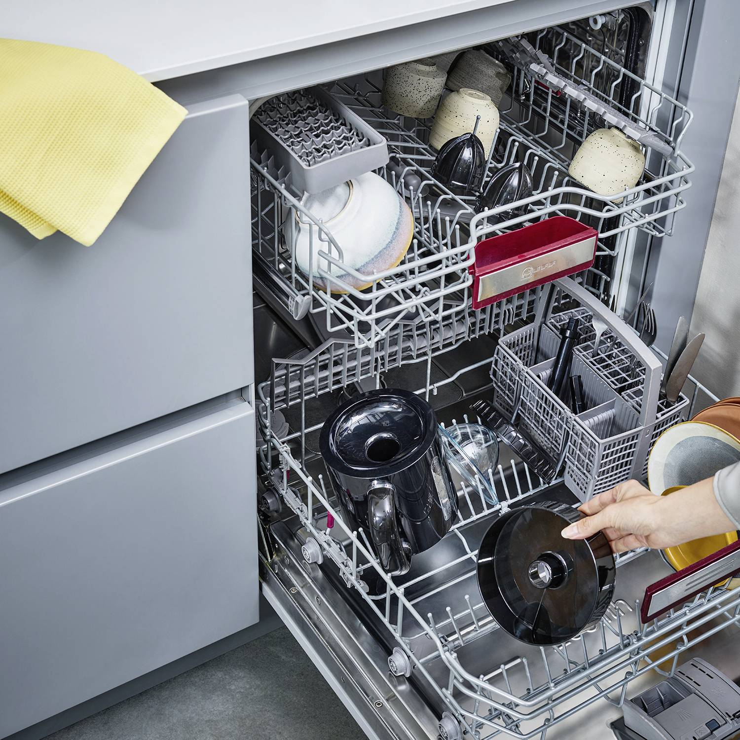 An open dishwasher full of crockery, including mugs, plates and cutlery, neatly arranged in racks. A hand is holding the bottom rack.