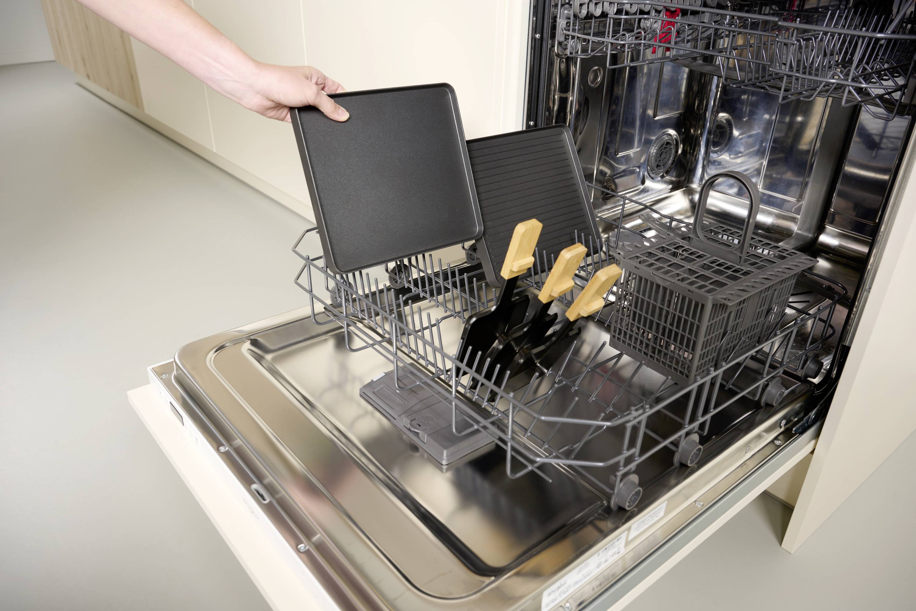 A hand places a baking tray into the open dishwasher. Inside the dishwasher are knives with orange handles.