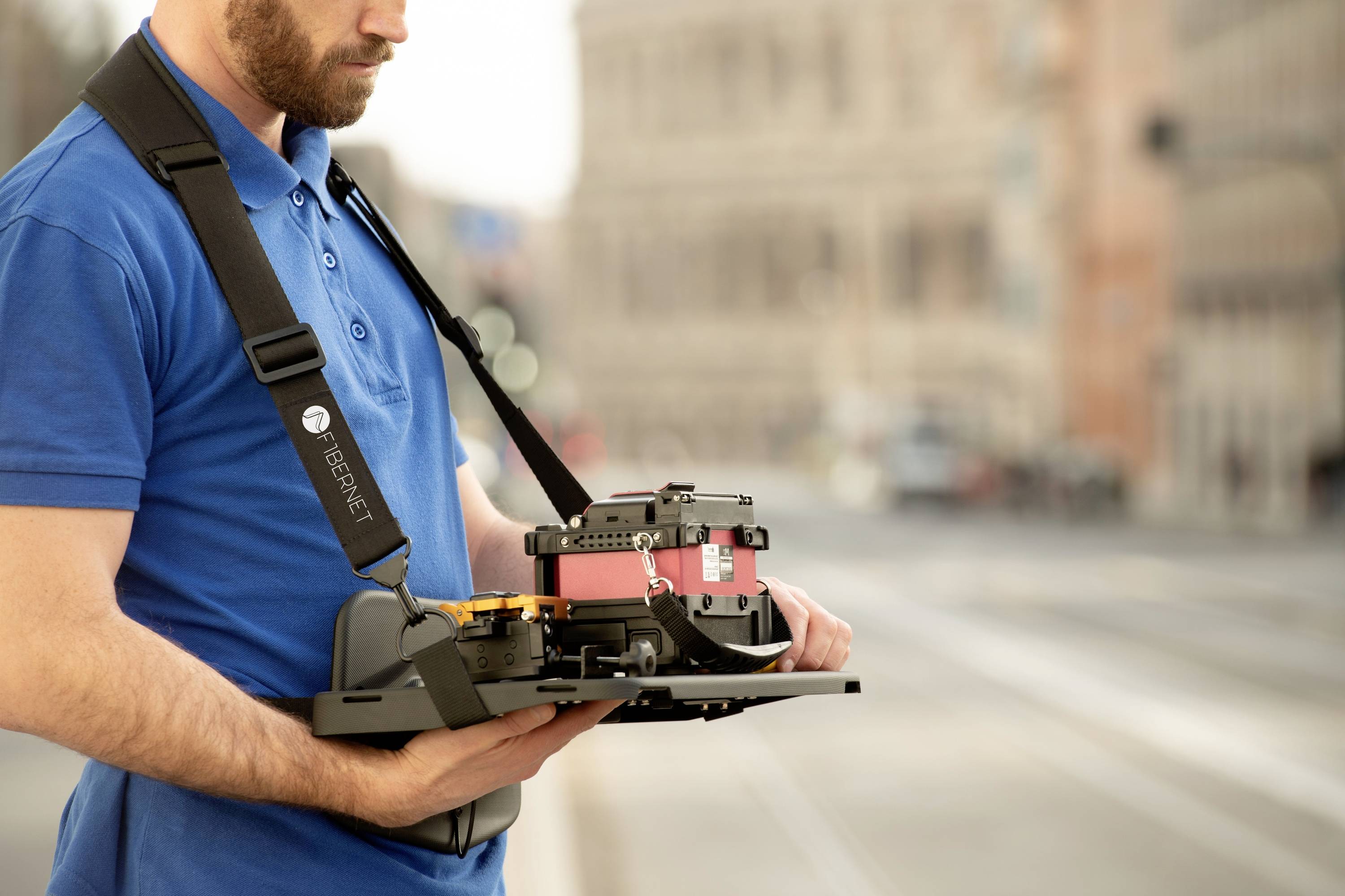 A man in a blue shirt is wearing a laser scanning device on a strap. A blurry urban environment can be seen in the background.