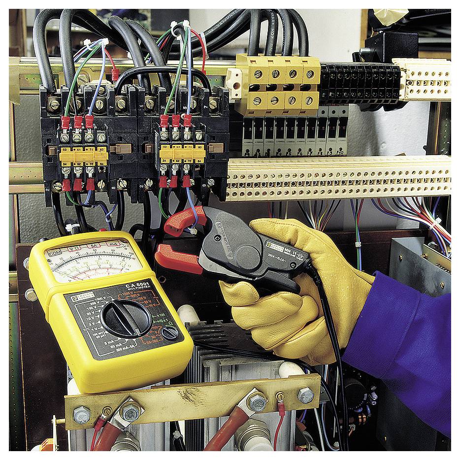 Someone wearing a yellow glove is checking electrical connections on a switchboard using a multimeter.
