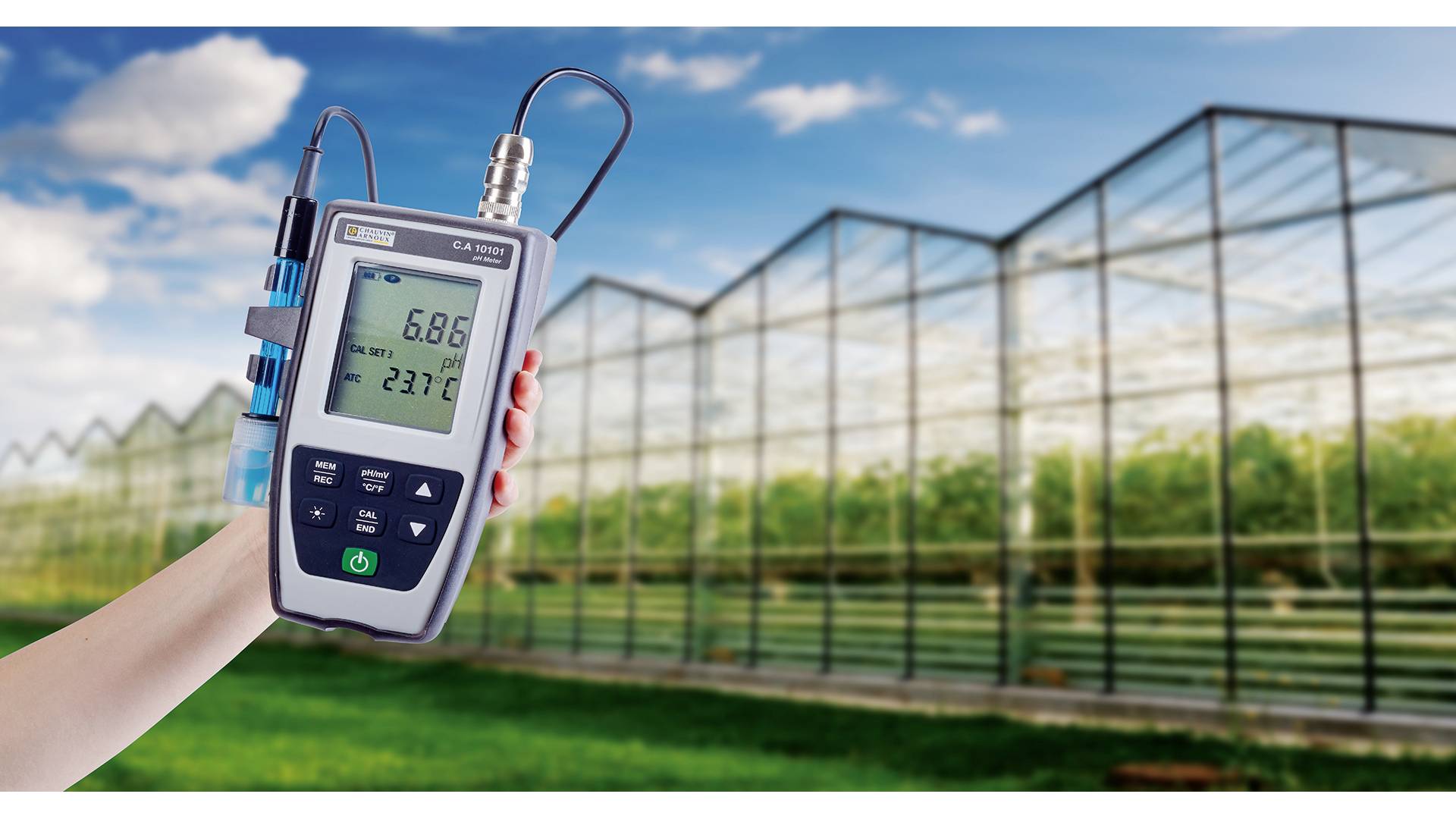 A portable measuring device with a display shows '6.86' and '23.7°C', held in front of a greenhouse.