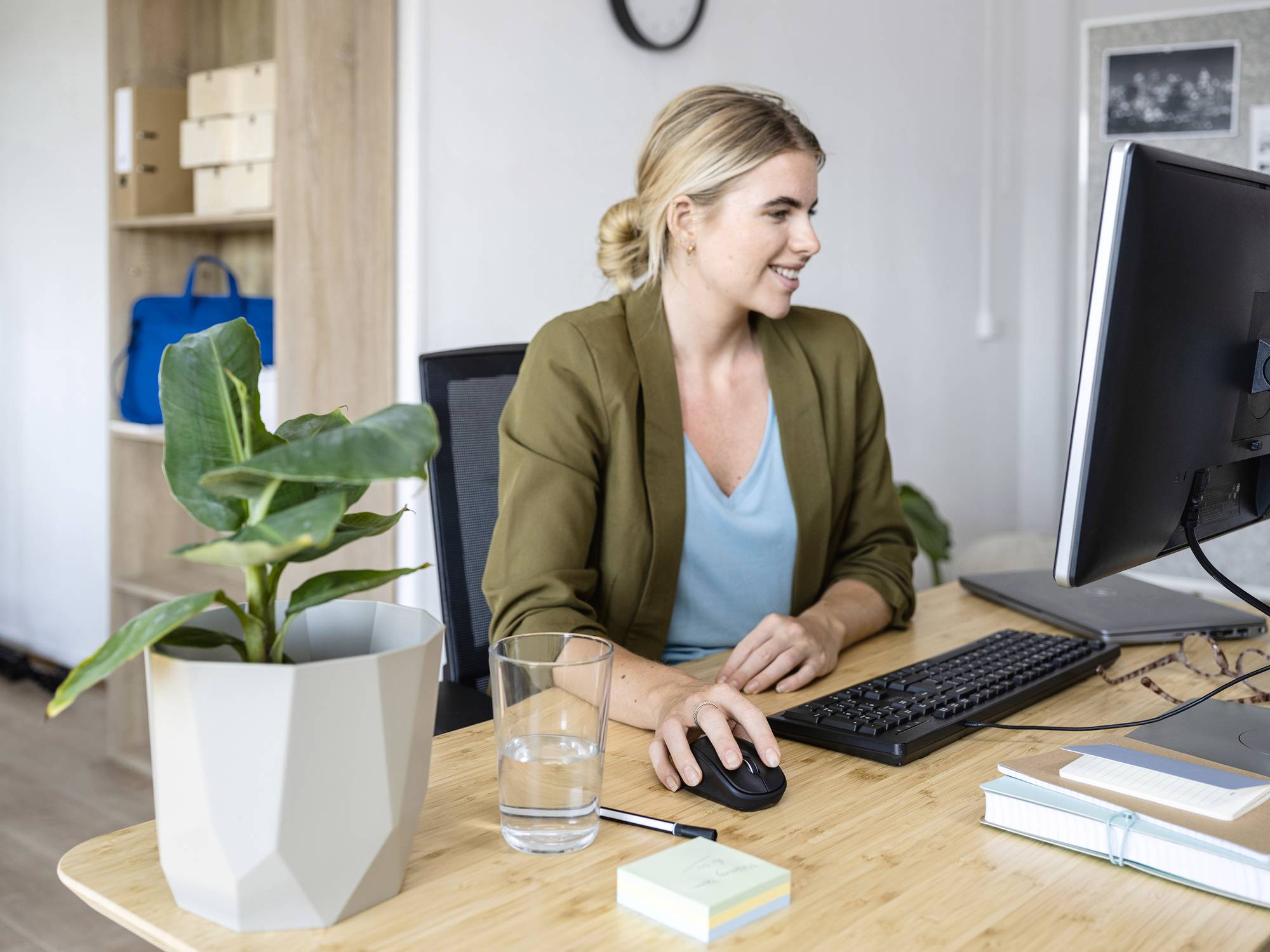 A woman sits smiling at her desk, working on a computer. A plant and a glass of water are beside her. Shelves can be seen in the background.