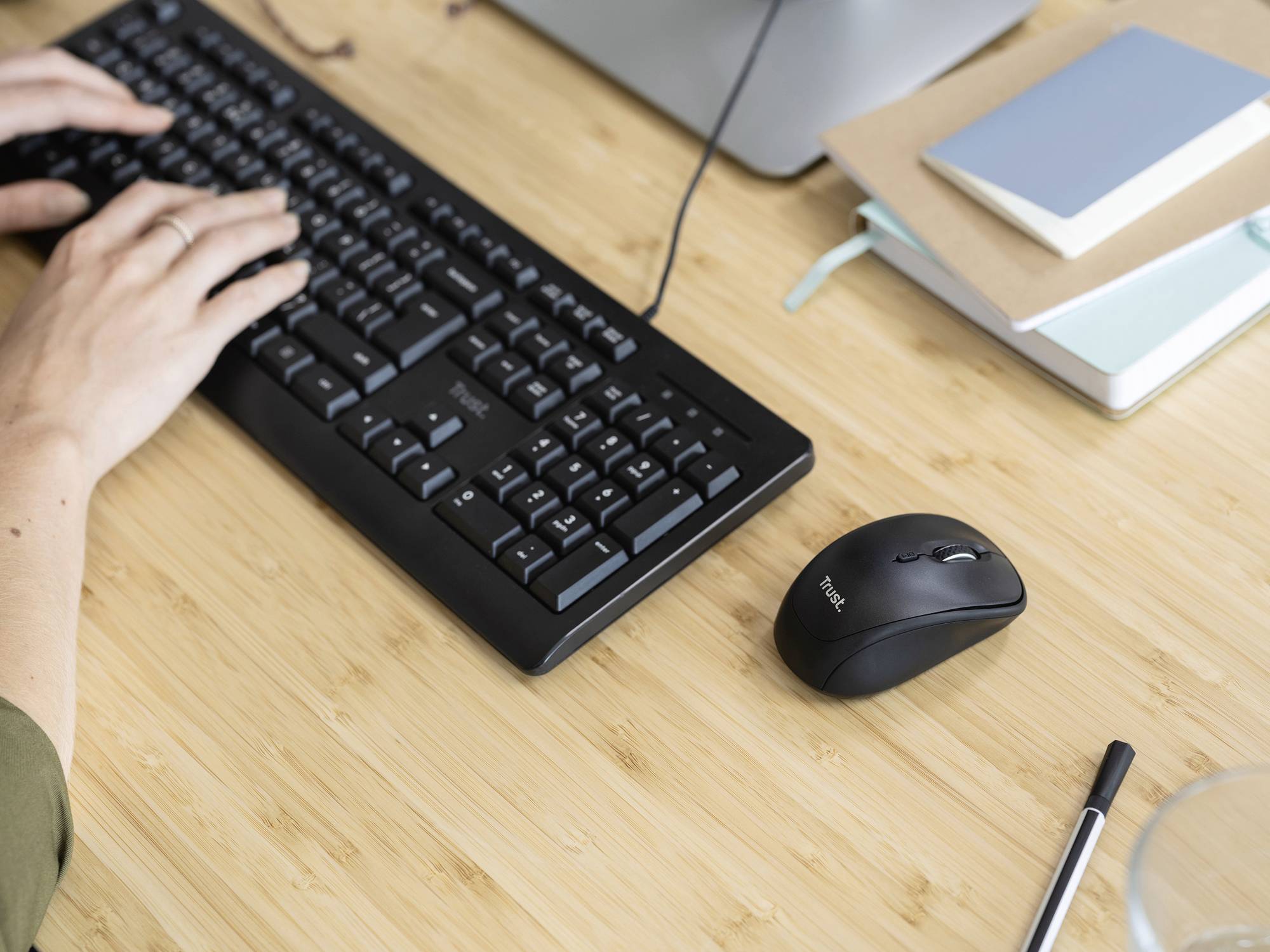 Hands typing on a keyboard on a desk with a mouse, pens, and notebooks nearby.