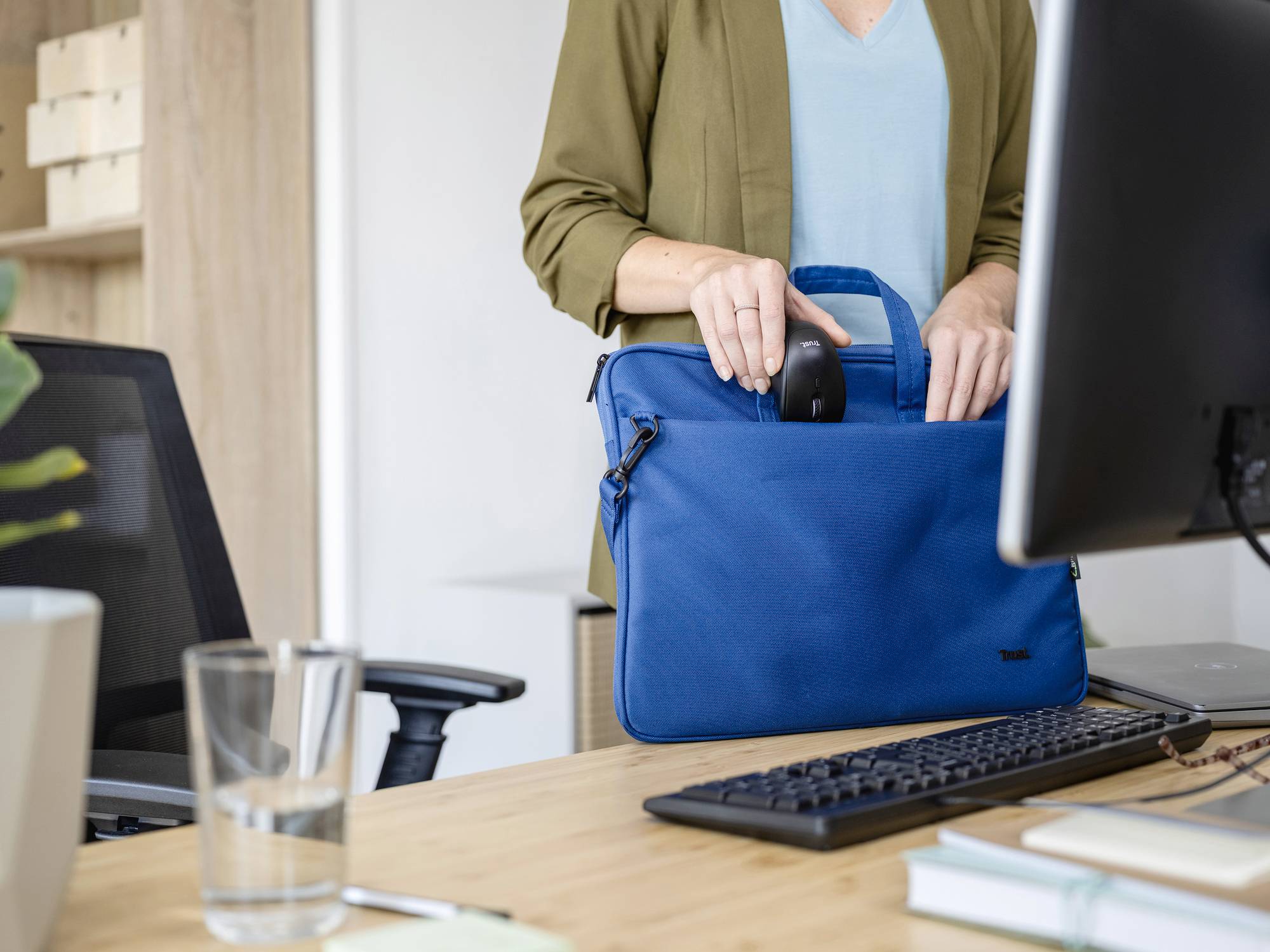 A person is storing a mouse in a blue laptop bag on a desk that also has a monitor, keyboard, and a glass on it. Office environment.