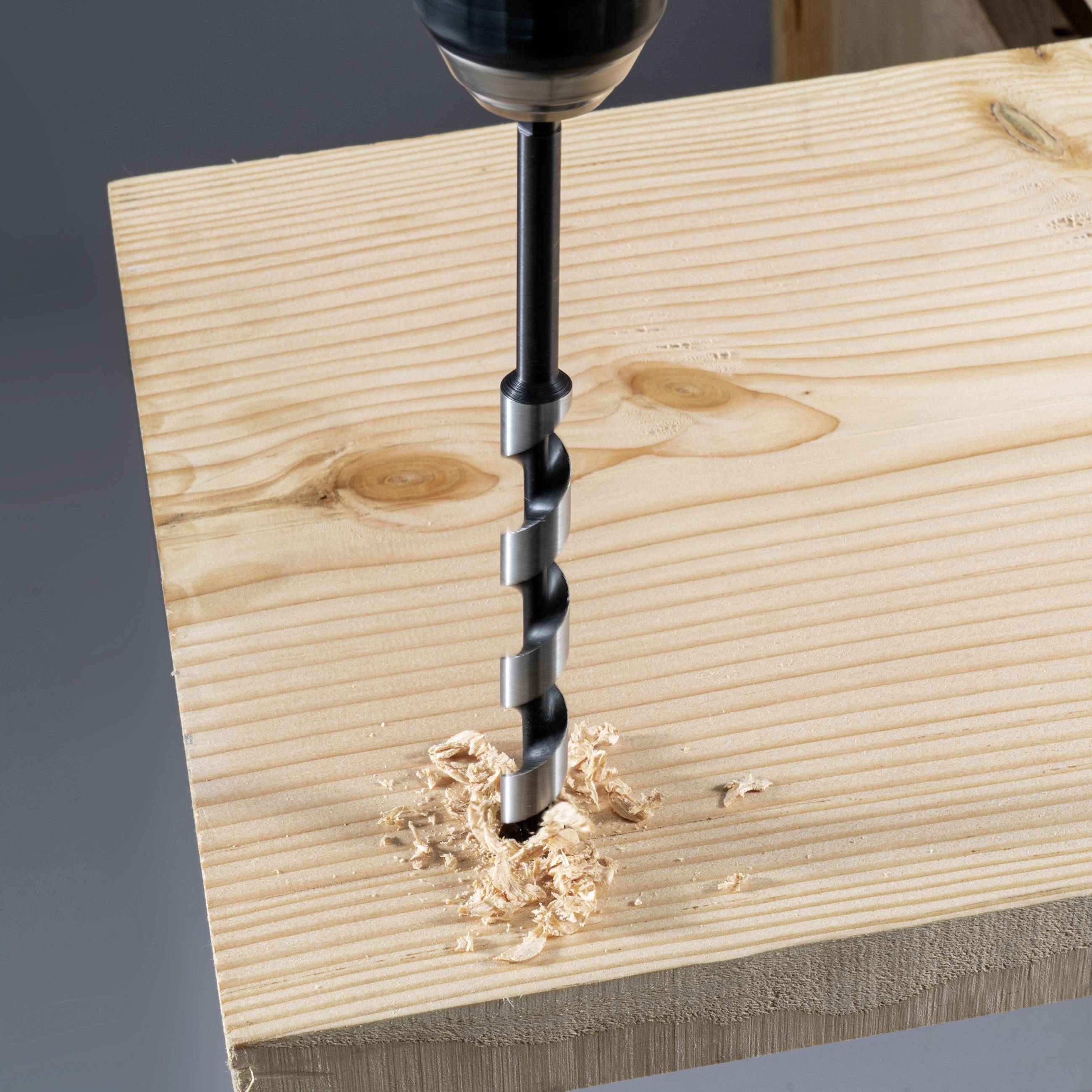 A drill bit pierces a wooden board, wood shavings collecting around the hole. The focus is on the drill tip and the severed wood.