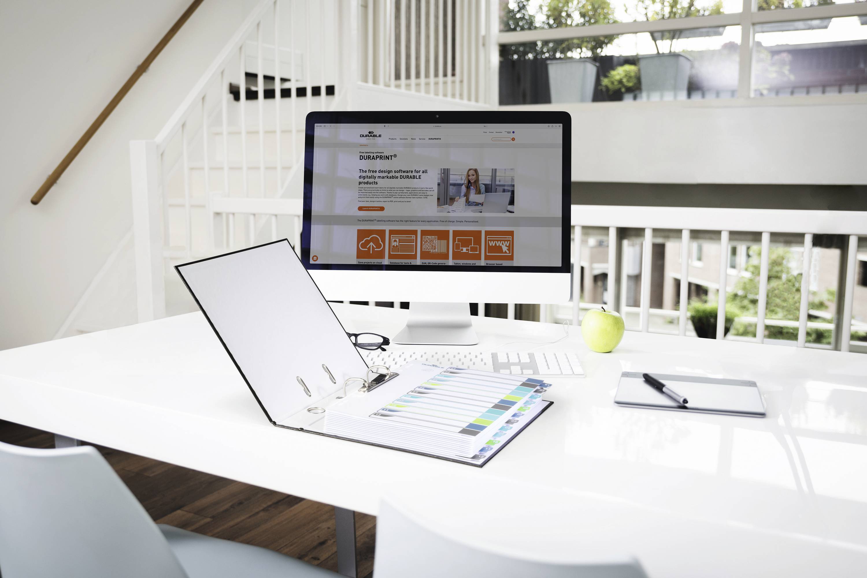 Modern workspace with a computer, notebook, colour palette and apple on a white desk in a bright room with large windows.