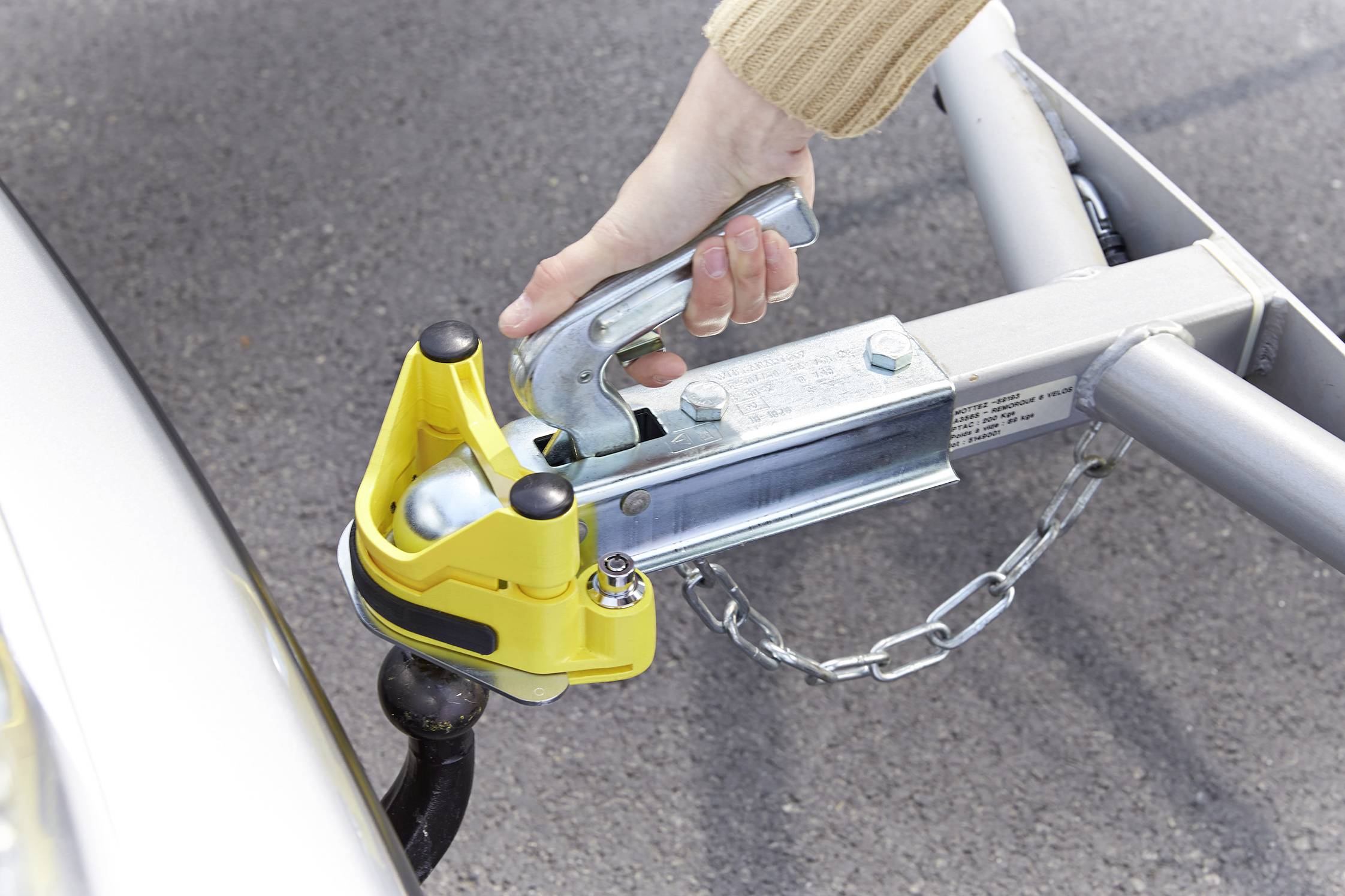 A hand is mounting a towbar onto a car. A yellow safety mechanism protects the connection.